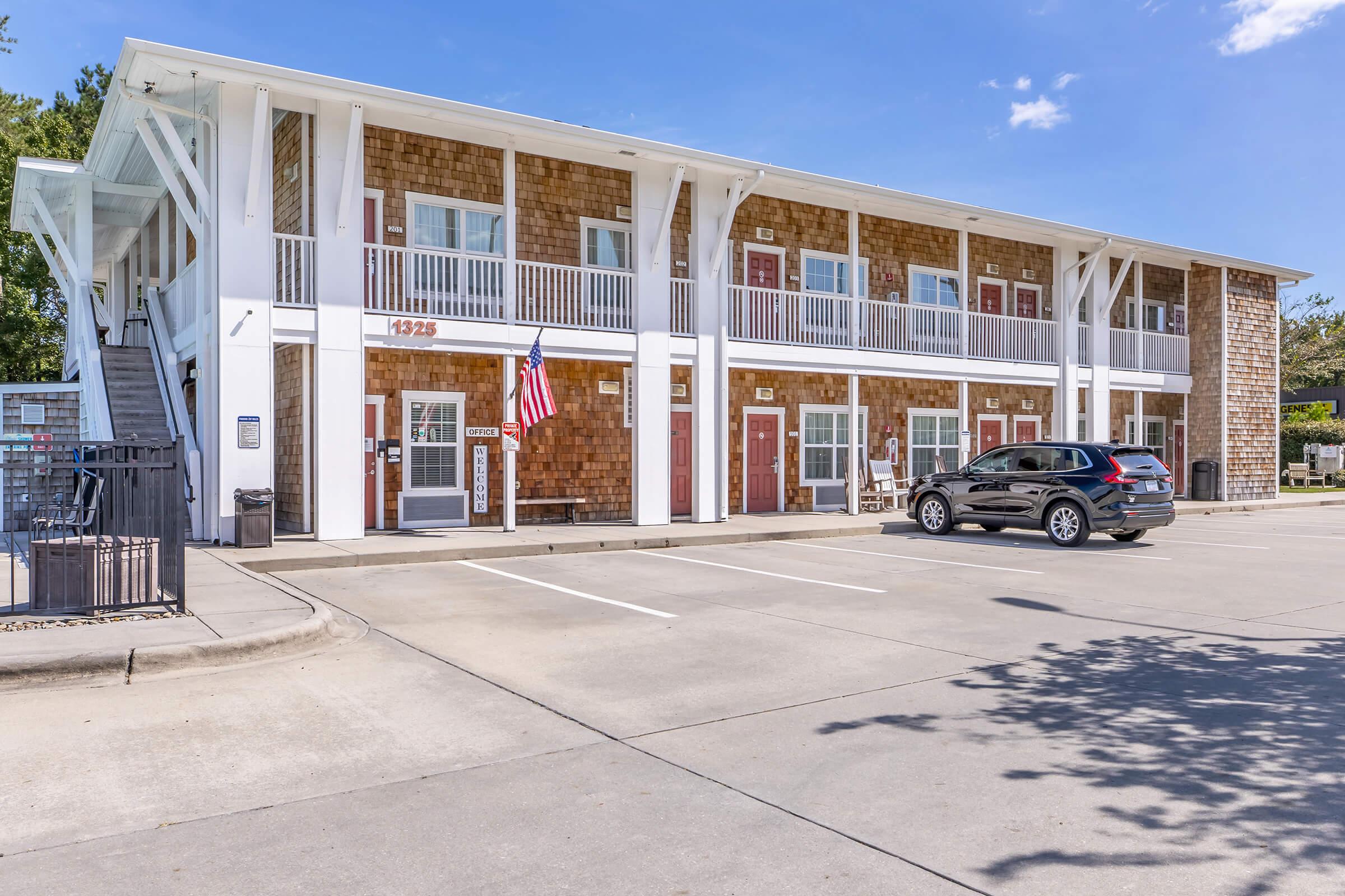 A multi-story building with a mix of wooden and white exterior. It features balconies, red doors, and a small American flag. There are parking spaces in front, with an SUV parked nearby. The scene is set under a clear blue sky with a few clouds.