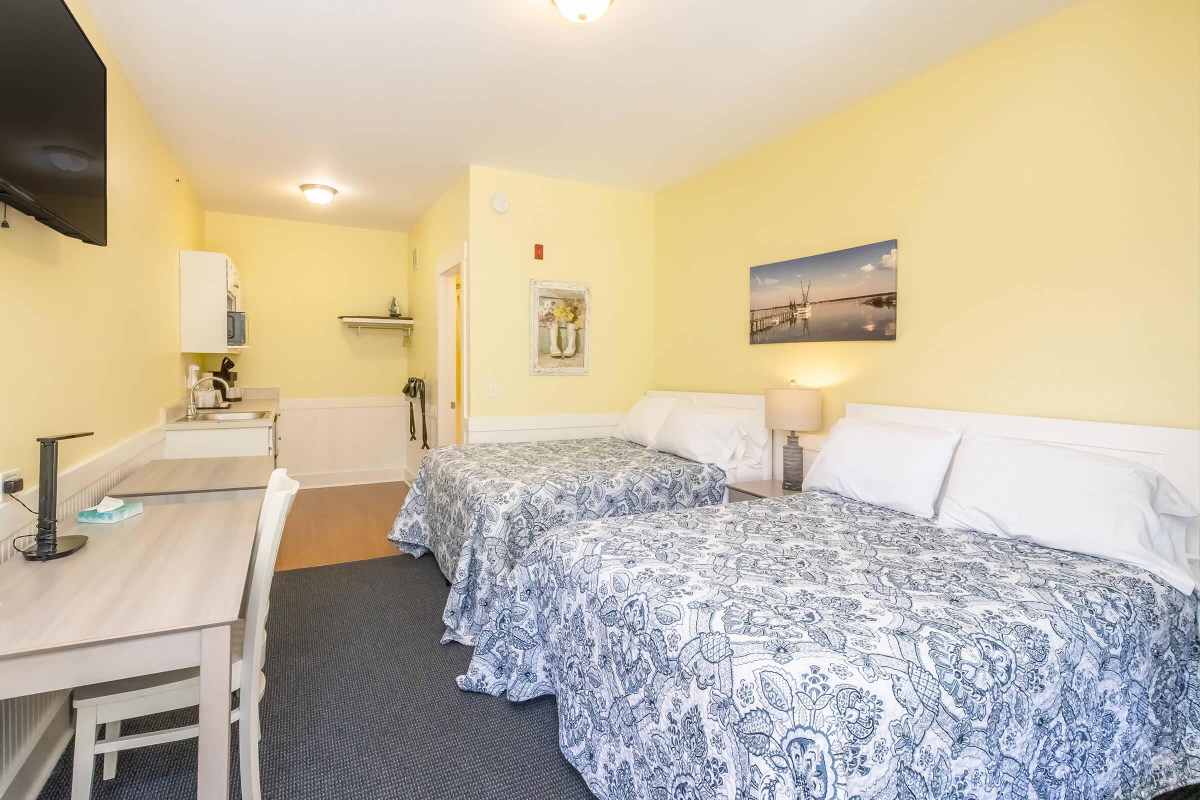 A cozy hotel room featuring two double beds with blue patterned bedspreads, a small table with a chair, a kitchenette area with a microwave and coffee maker, and a flat-screen TV mounted on the wall. The walls are painted a soft yellow, giving the room a warm and inviting feel. A framed picture of a waterfront scene is hung above the beds.