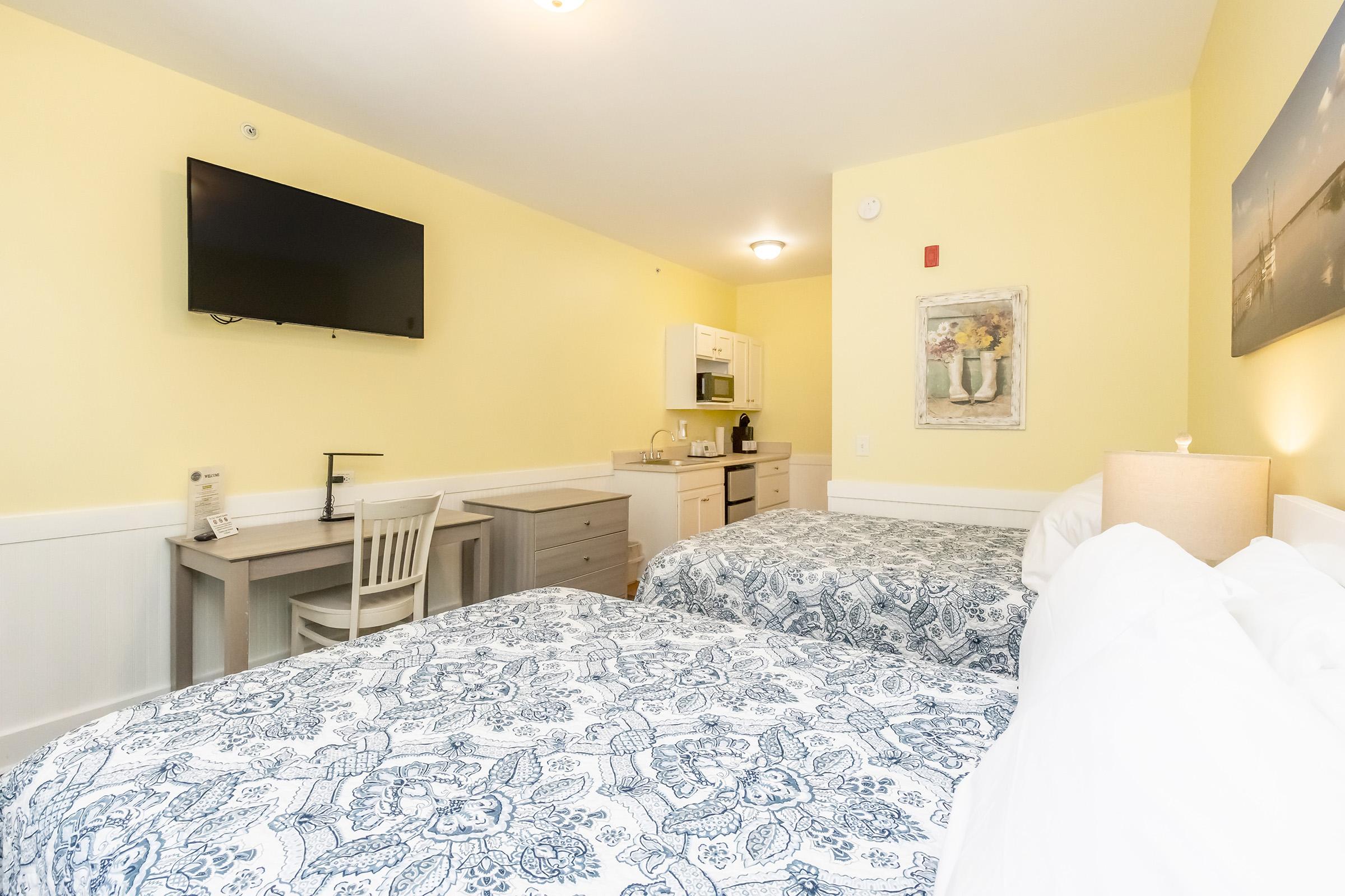 A brightly lit hotel room featuring two beds with blue and white patterned bedding. There's a small kitchenette with a microwave and a dining table. A flat-screen TV is mounted on the wall, and the room is painted in a cheerful yellow shade, creating a welcoming atmosphere.