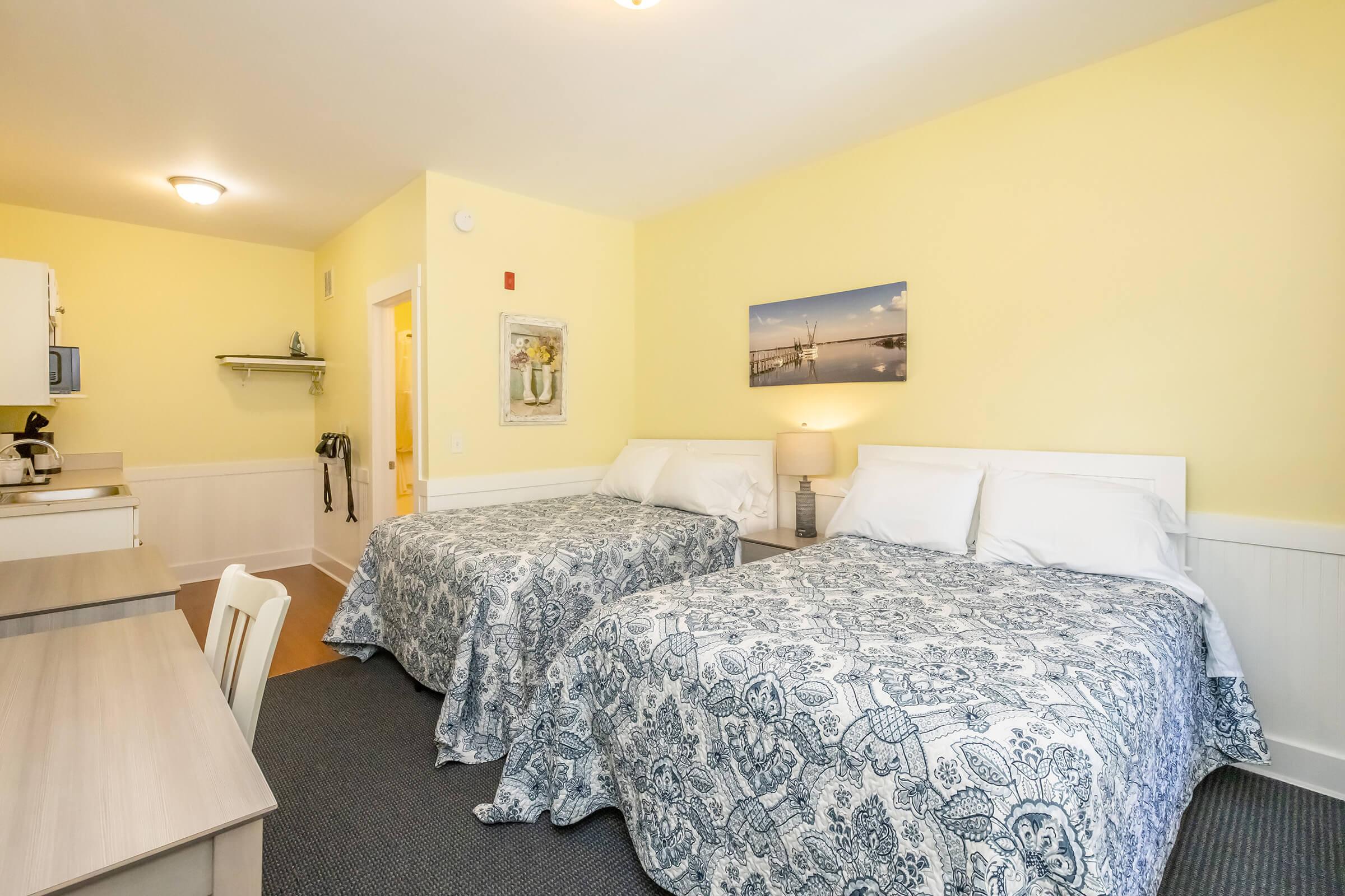 A cozy hotel room featuring two double beds with blue and white patterned bedspreads, a small dining table with chairs, and a kitchenette area. The walls are painted a warm yellow, and there is a picture of a serene landscape mounted above the beds. The space is well-lit and inviting.