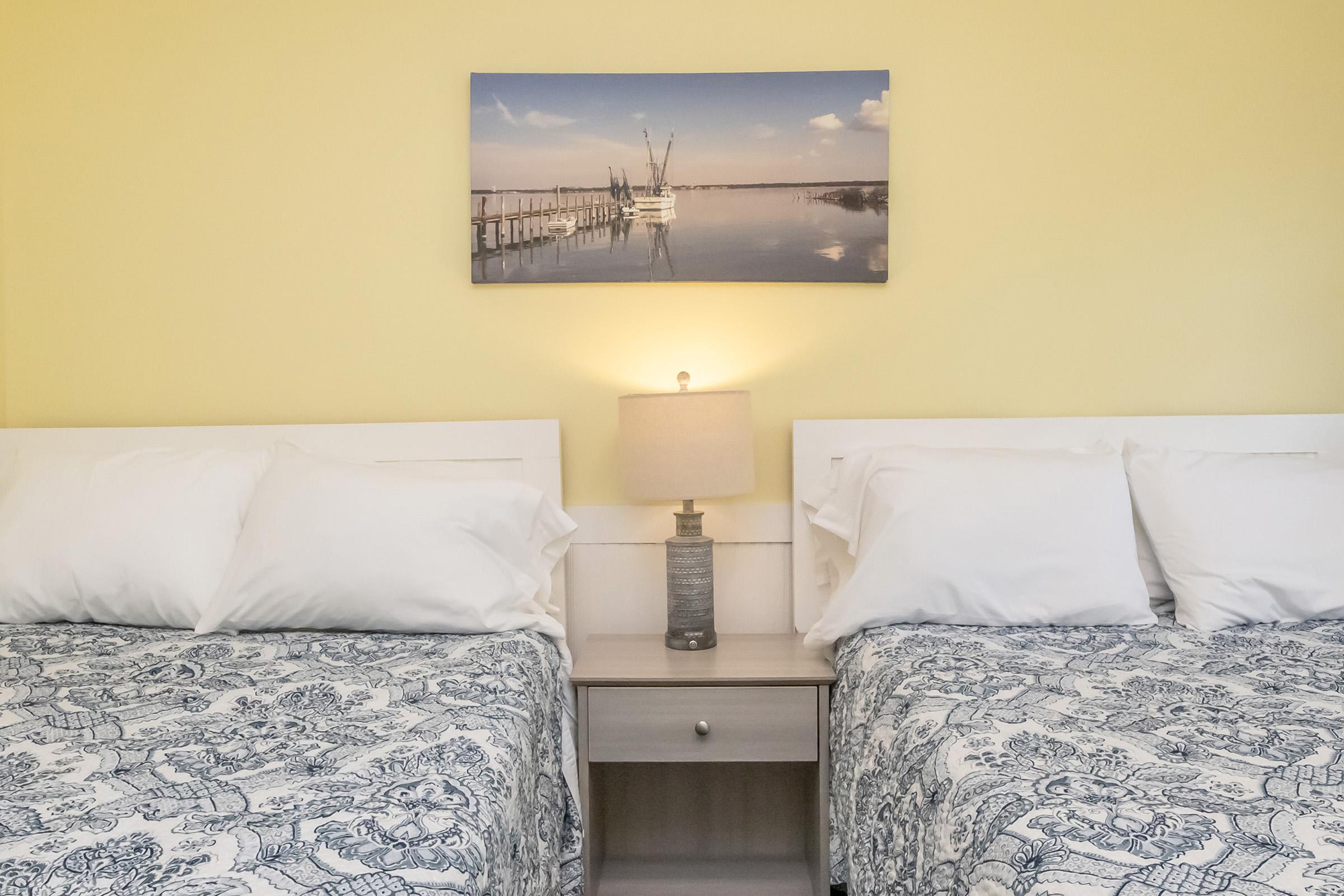 A cozy bedroom with two neatly made beds featuring patterned blue and white bedspreads. Between the beds is a small nightstand with a lamp. Above the beds, a landscape photograph of a serene waterfront scene hangs on the yellow wall, contributing to a warm atmosphere.