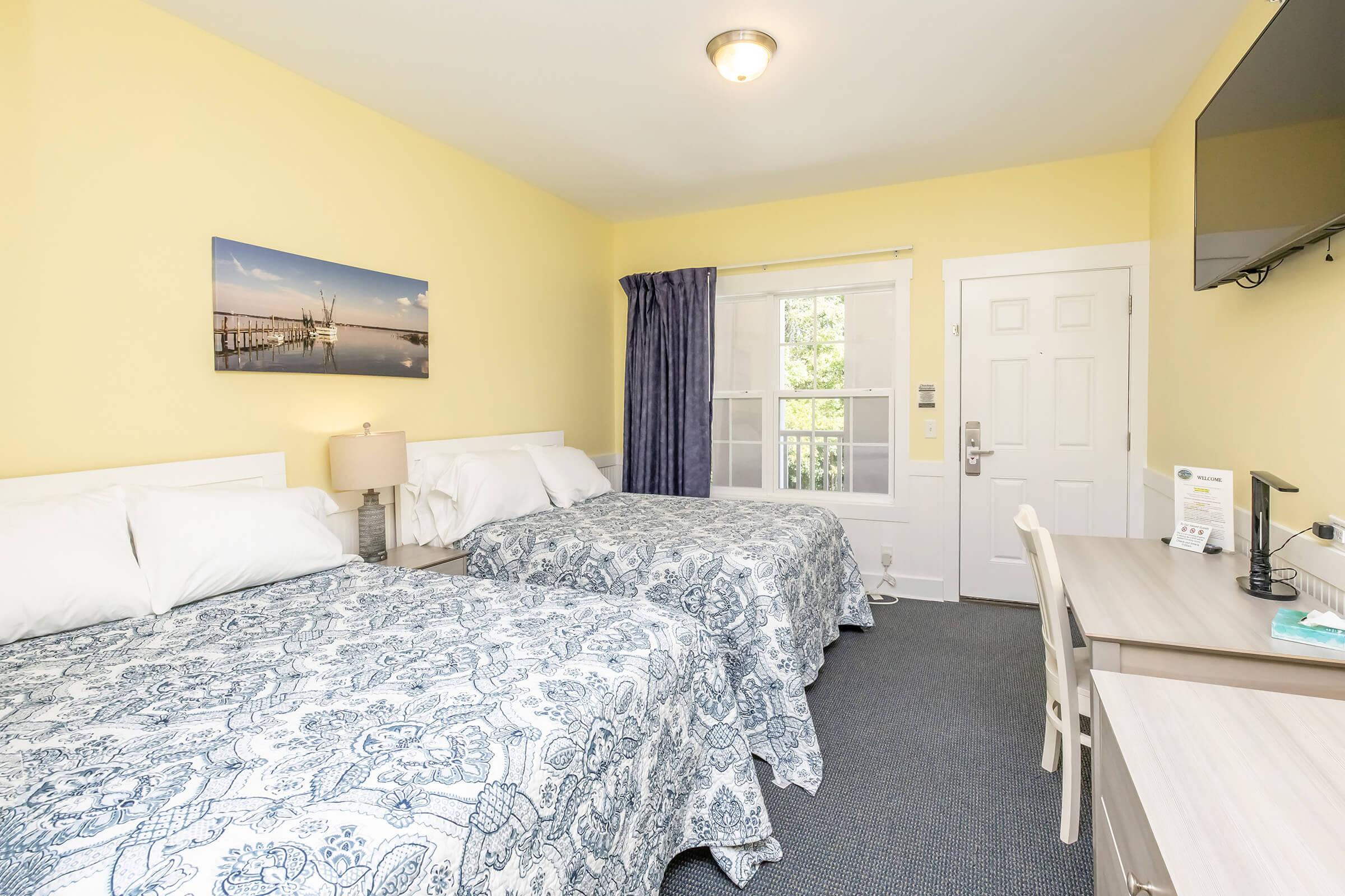 A cozy hotel room featuring two double beds with blue and white patterned bedspreads. There's a light yellow wall, a desk with a chair, and a television mounted on the wall. A window with a blue curtain offers natural light, and a scenic picture of a pier hangs above the bed.