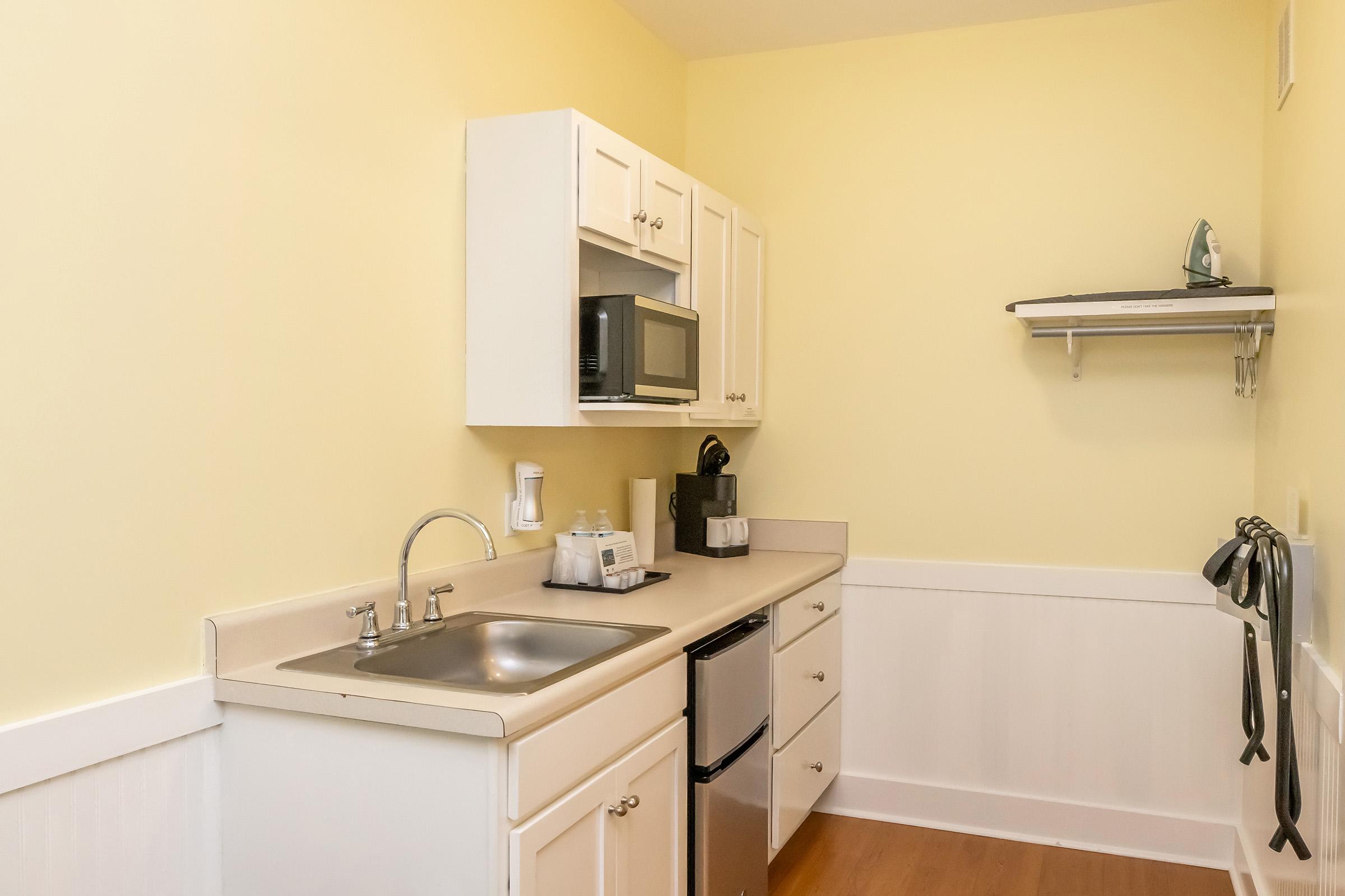 A small kitchen area featuring a yellow wall, white cabinets, a stainless steel sink, a microwave, and a compact refrigerator. A coffee maker is on the counter beside a set of cups. There is a shelf above the counter, and kitchen utensils are hanging on the wall, with wooden flooring completing the space.
