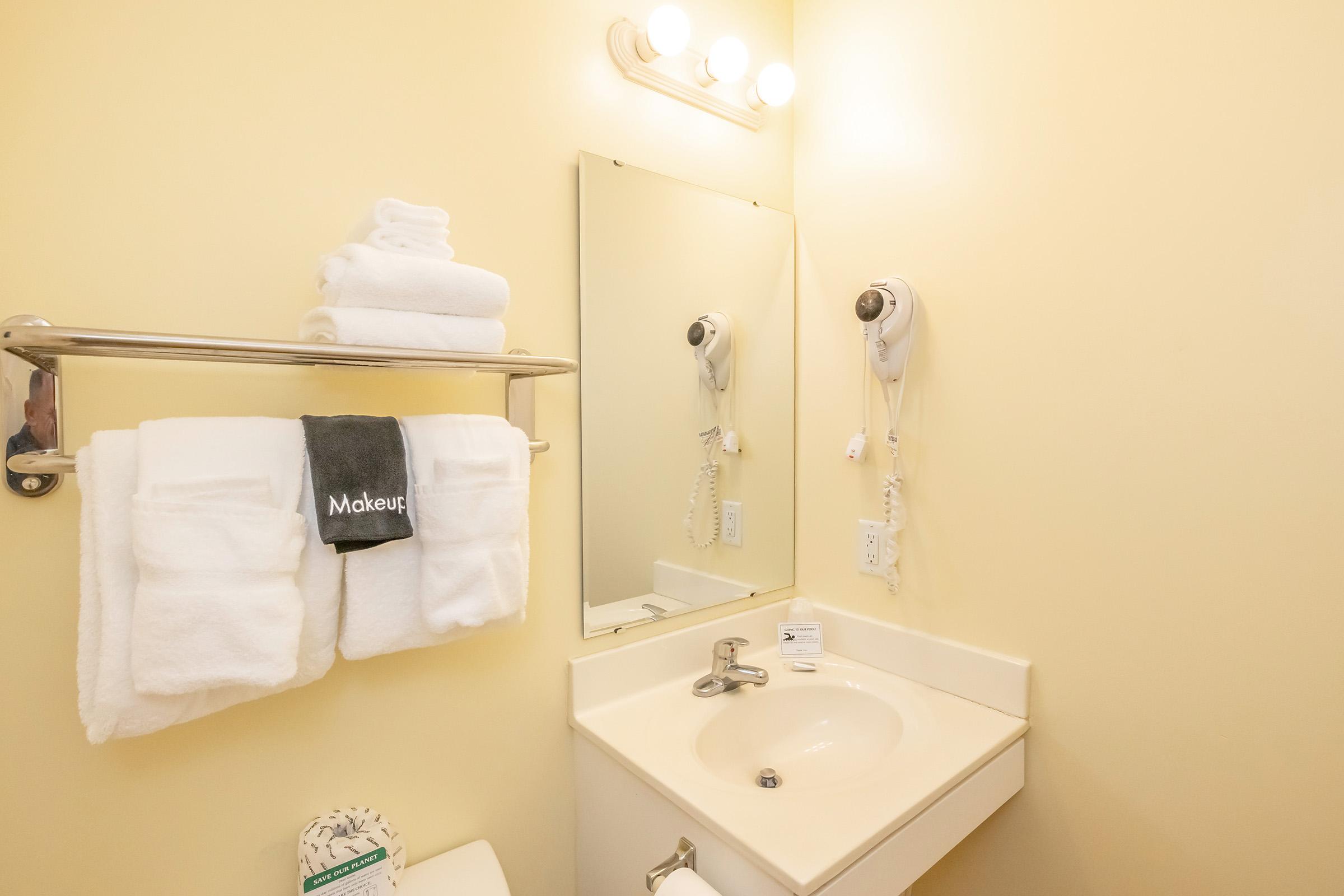 A small bathroom featuring a sink with a mirror above, a hand towel rack holding neatly folded white towels, and a wall-mounted hairdryer. The walls are painted yellow, and there is a light fixture above the mirror. A roll of toilet paper is visible, along with an informational sign.