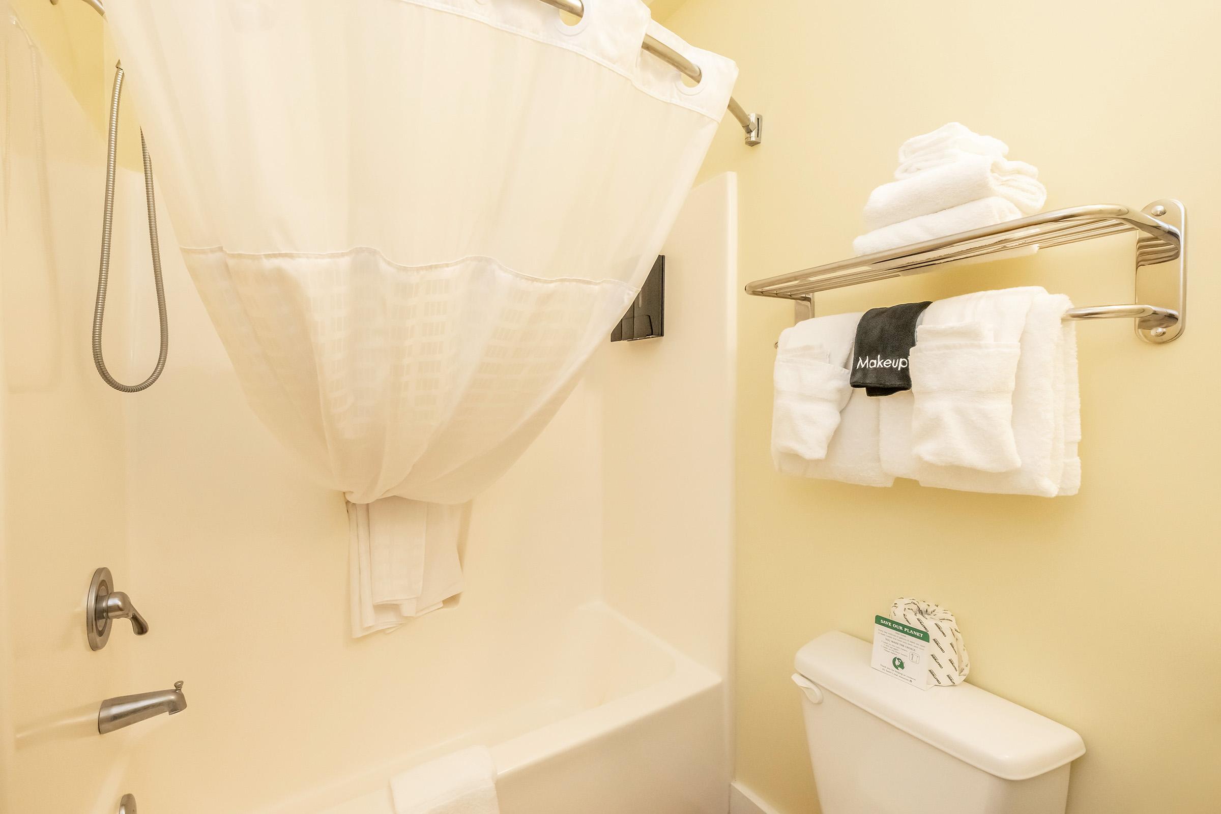 A small bathroom featuring a shower with a white curtain, neatly folded white towels on a rack, and a white toilet. The walls are painted a light yellow, creating a bright and clean atmosphere. A bar of soap is displayed on the toilet tank.