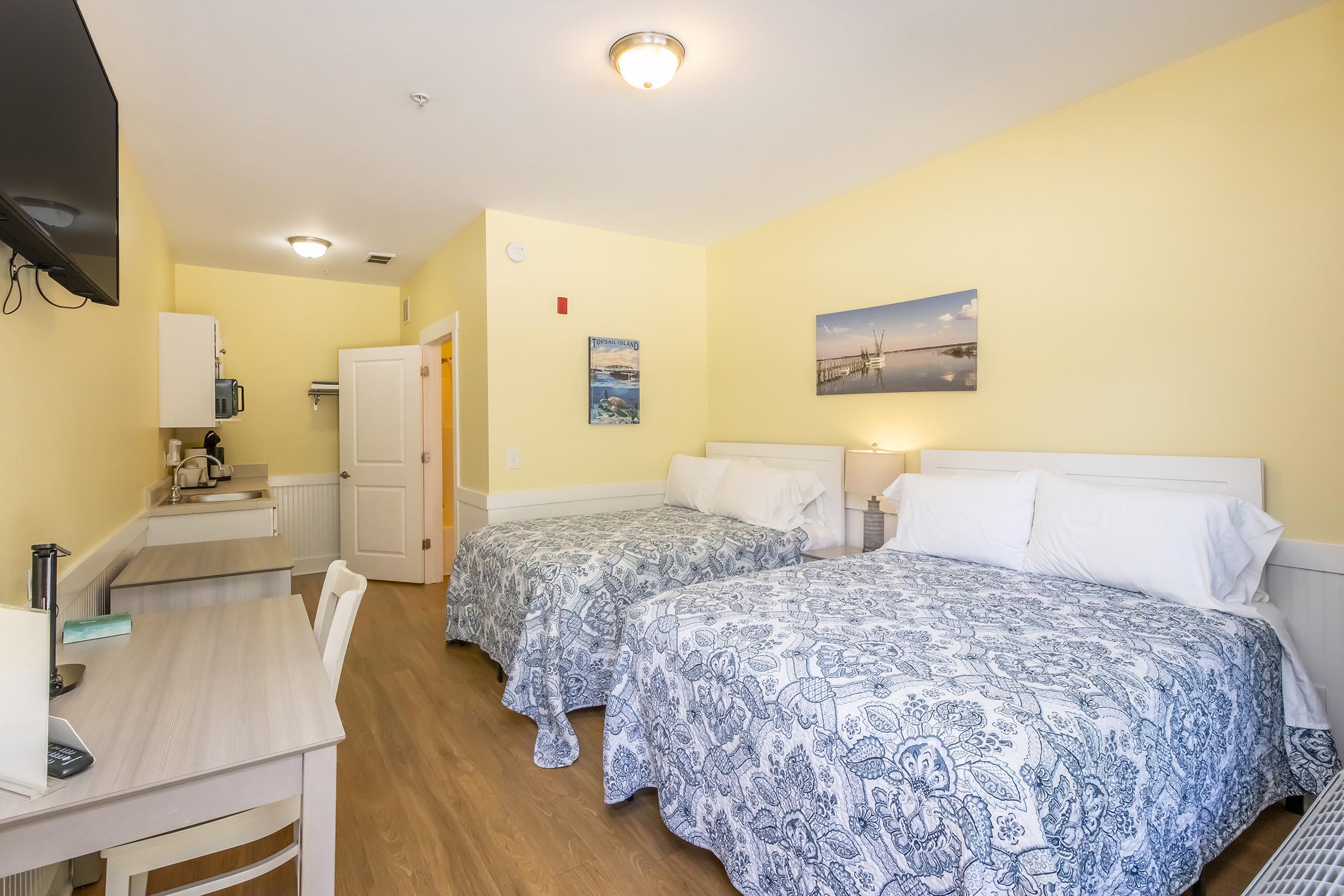 A cozy hotel room featuring two beds with blue and white patterned comforters. The walls are painted yellow, and there's a flat-screen TV mounted on the wall. A small workspace with a desk and chair is present, along with a kitchenette area. Soft lighting creates a welcoming atmosphere.