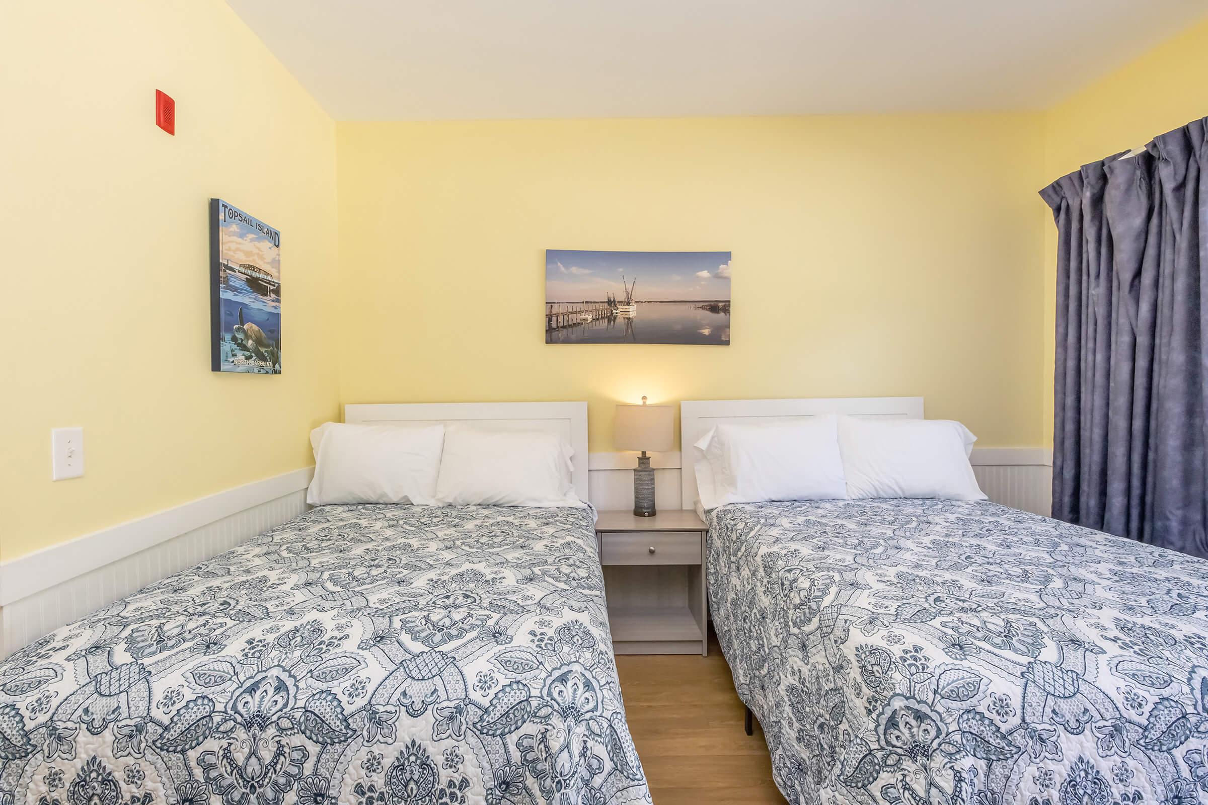A cozy hotel room featuring two neatly made beds with patterned bedspreads, a bedside table with a lamp, and soft yellow walls. A photograph of a serene pier scene hangs on the wall, complemented by a curtain covering a window. A pop of color is added with a red wall-mounted device.