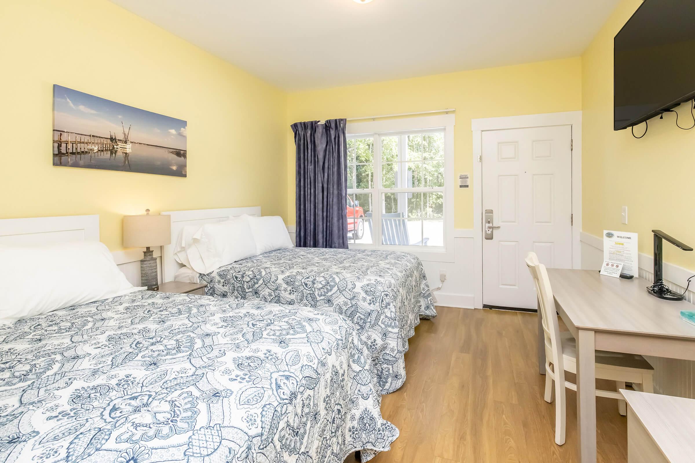 A bright and airy hotel room featuring two beds with blue patterned bedspreads. The walls are painted yellow, and a large window lets in natural light. A small desk and chair are positioned next to the window, and a flat-screen TV is mounted on the wall. A door leads outside, enhancing the room's inviting atmosphere.