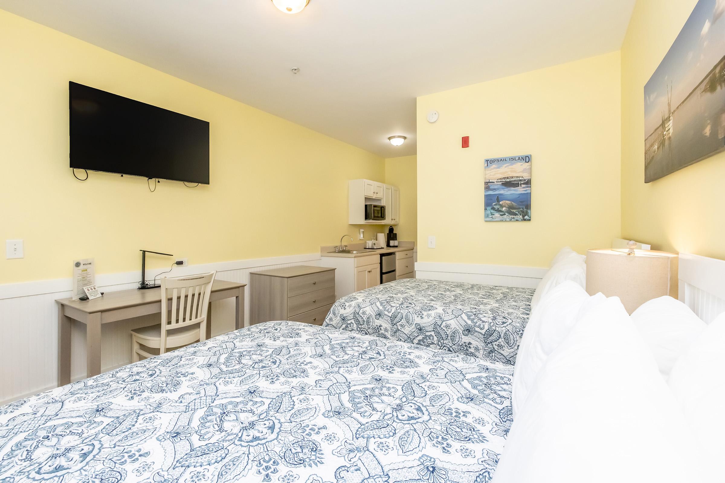 A cozy hotel room featuring two double beds with blue patterned bedspreads, a kitchenette with a microwave and mini-fridge, a desk with a chair, and a television mounted on the wall. The walls are painted a soft yellow, creating a warm atmosphere. There's a piece of artwork hanging above the beds.