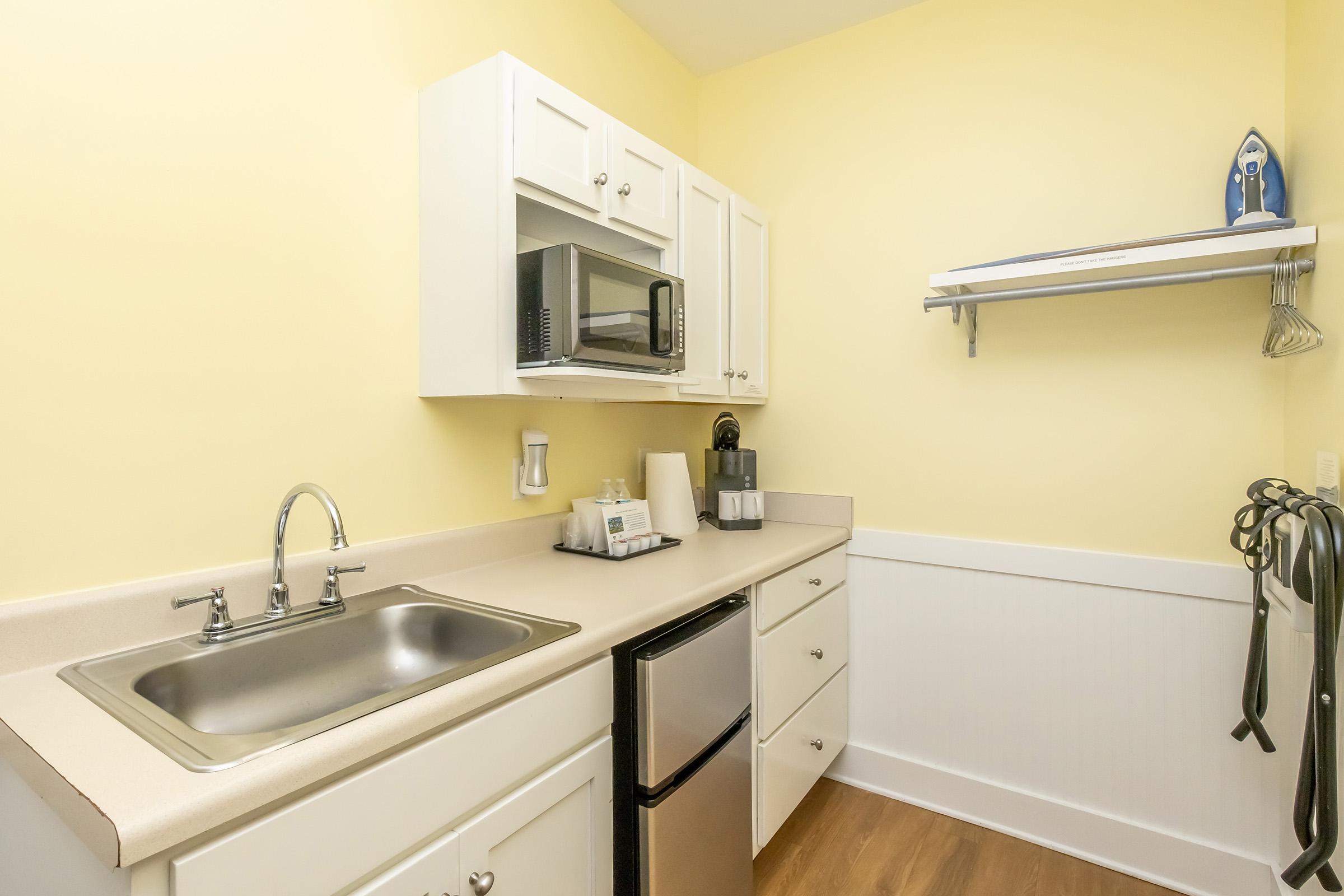 A compact kitchen featuring yellow walls, a stainless steel sink, a microwave, and a refrigerator. There are white cabinets above the countertop, and on the wall, there is a shelf holding an iron and hangers. The floor is a warm wood tone, contributing to a cozy atmosphere.