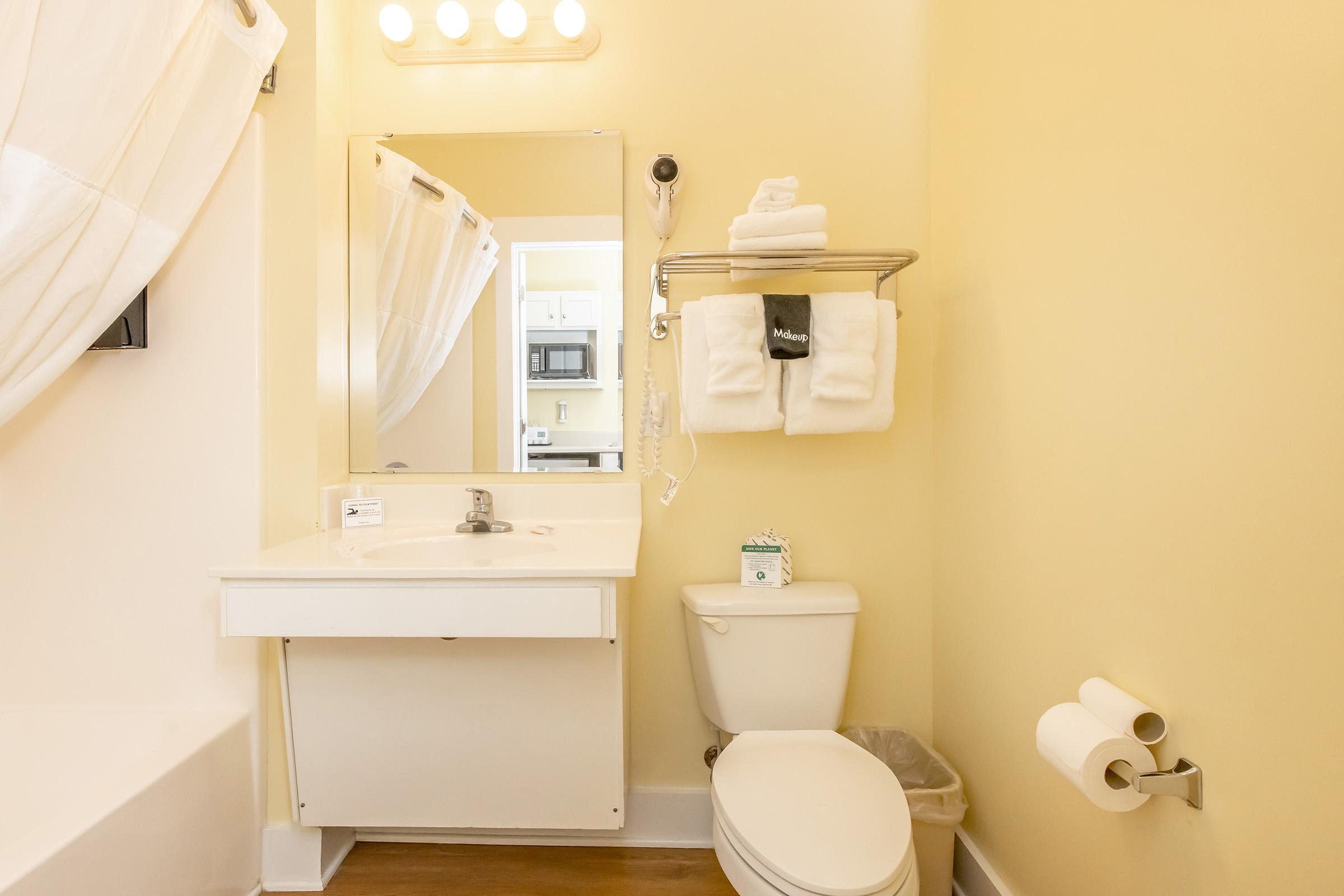 A clean bathroom featuring a white sink with a mirror above, a toilet, and a shower with a curtain. Towels are neatly stacked on a wall-mounted shelf, and there's a small trash bin. The walls are painted a soft yellow, creating a bright and airy atmosphere.