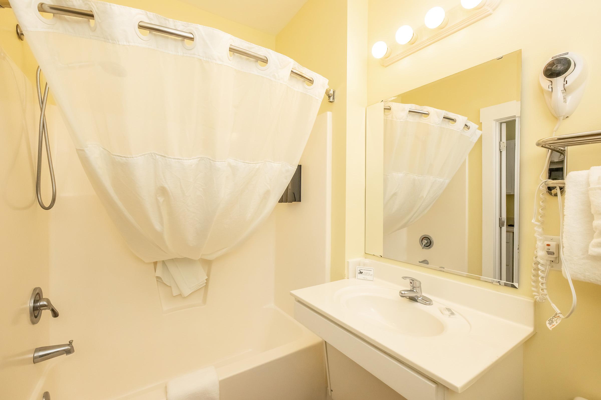 A clean and simple bathroom featuring a bathtub with a shower curtain, a sink with a mirror above, and a hairdryer mounted on the wall. The walls are painted a soft yellow, enhancing the bright and airy feel of the space. White towels are neatly arranged on a rack for convenience.