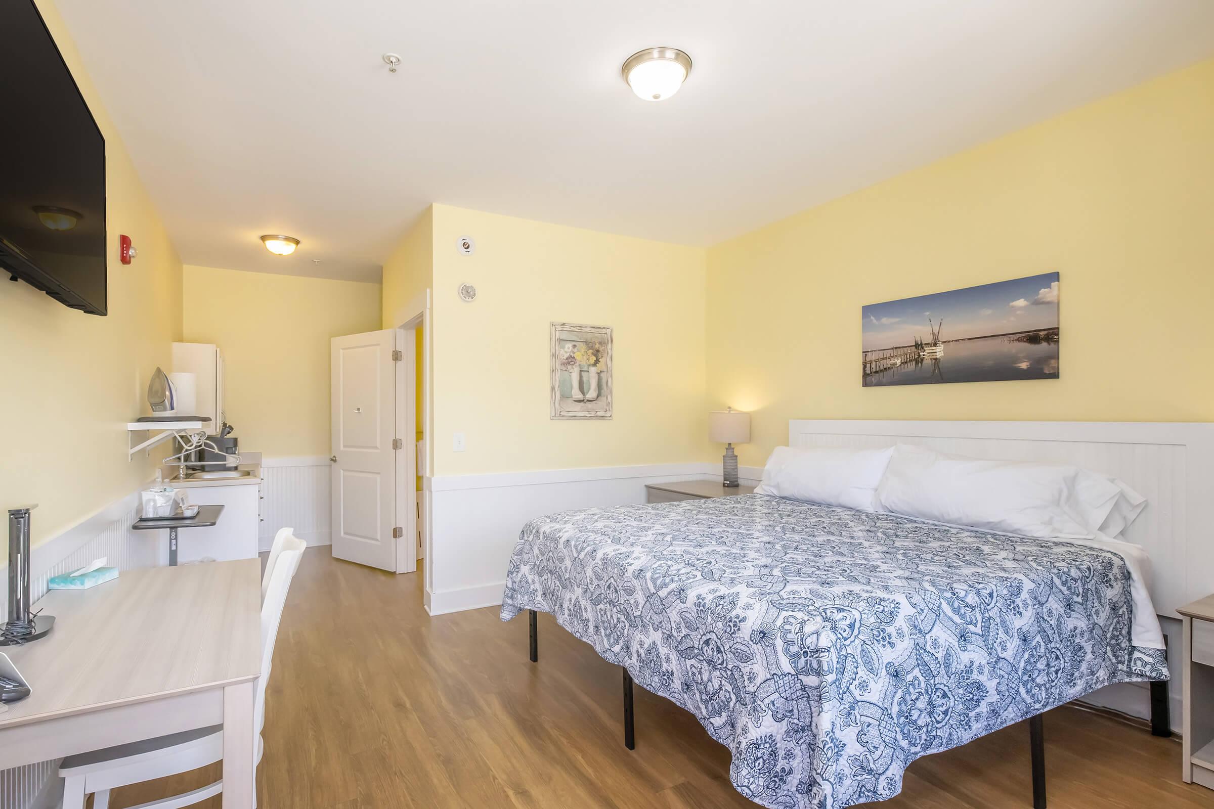 A brightly lit room featuring a large bed with a blue patterned bedspread. There is a small kitchenette area in the background, and a desk with a chair. A painting of a serene landscape hangs on the wall, and the walls are painted a soft yellow. A flat-screen TV is mounted on the wall opposite the bed.