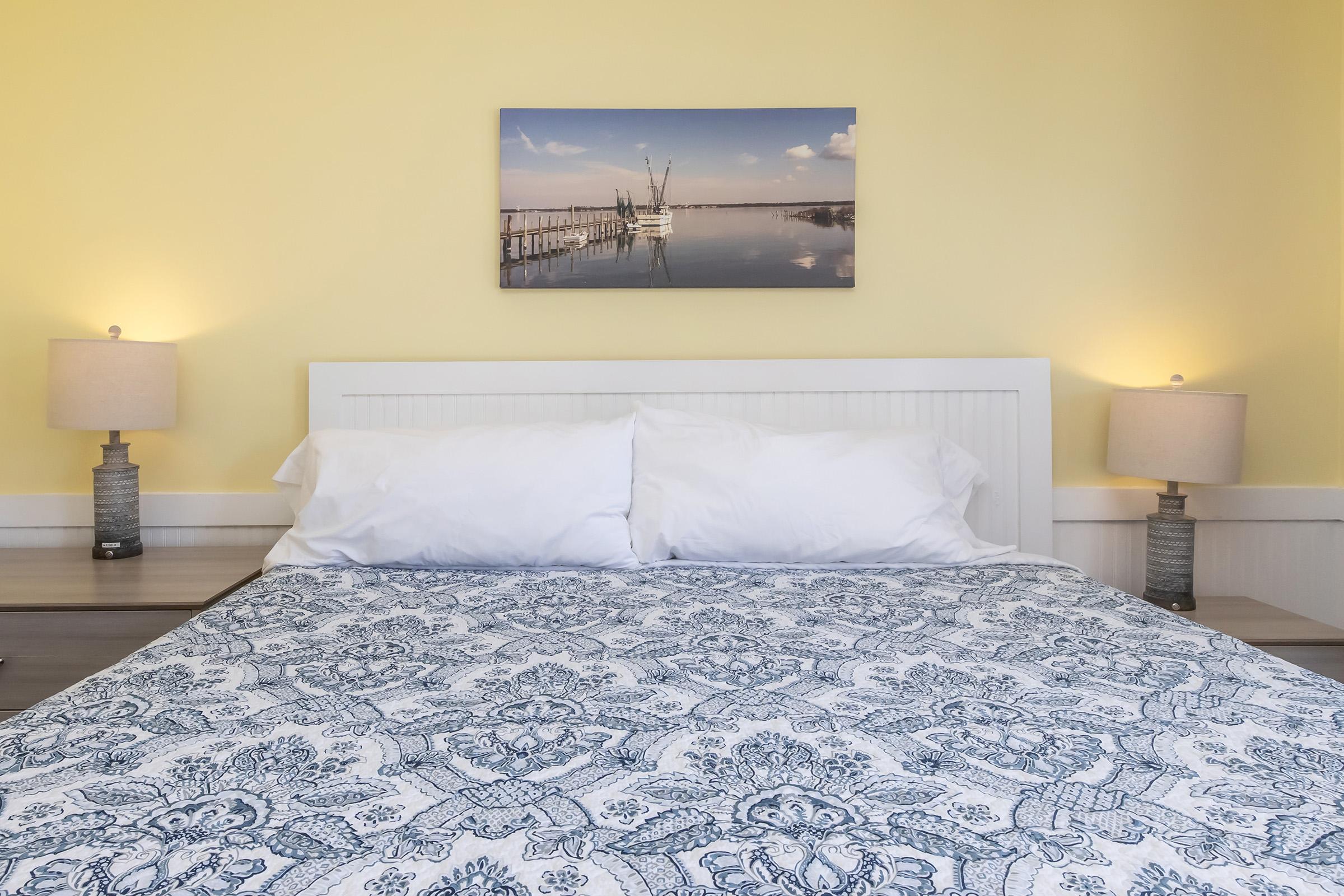 A cozy bedroom featuring a white bed with a blue and white patterned quilt. Two bedside lamps are on either side of the bed, and above it, there's a serene landscape painting of a dock reflecting in water, set against a light yellow wall.