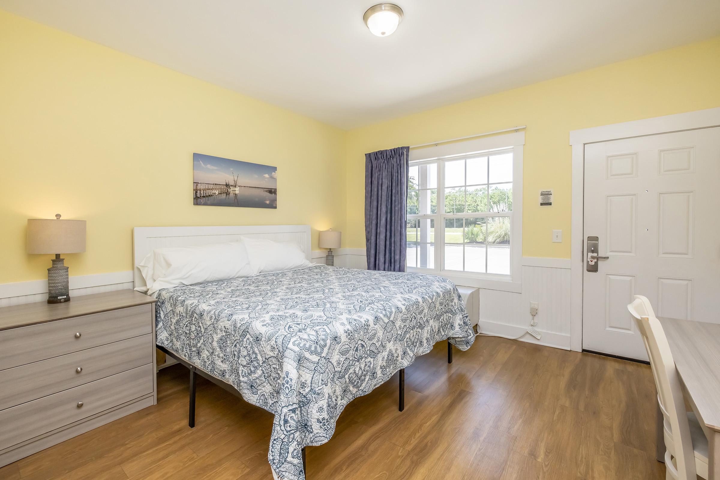 A cozy bedroom featuring a large bed with blue patterned bedding, a nightstand with a lamp, and a wooden dresser. There's a window with light-colored curtains allowing natural light, and wall decor showcasing a beach scene. The walls are painted yellow, creating a warm atmosphere.