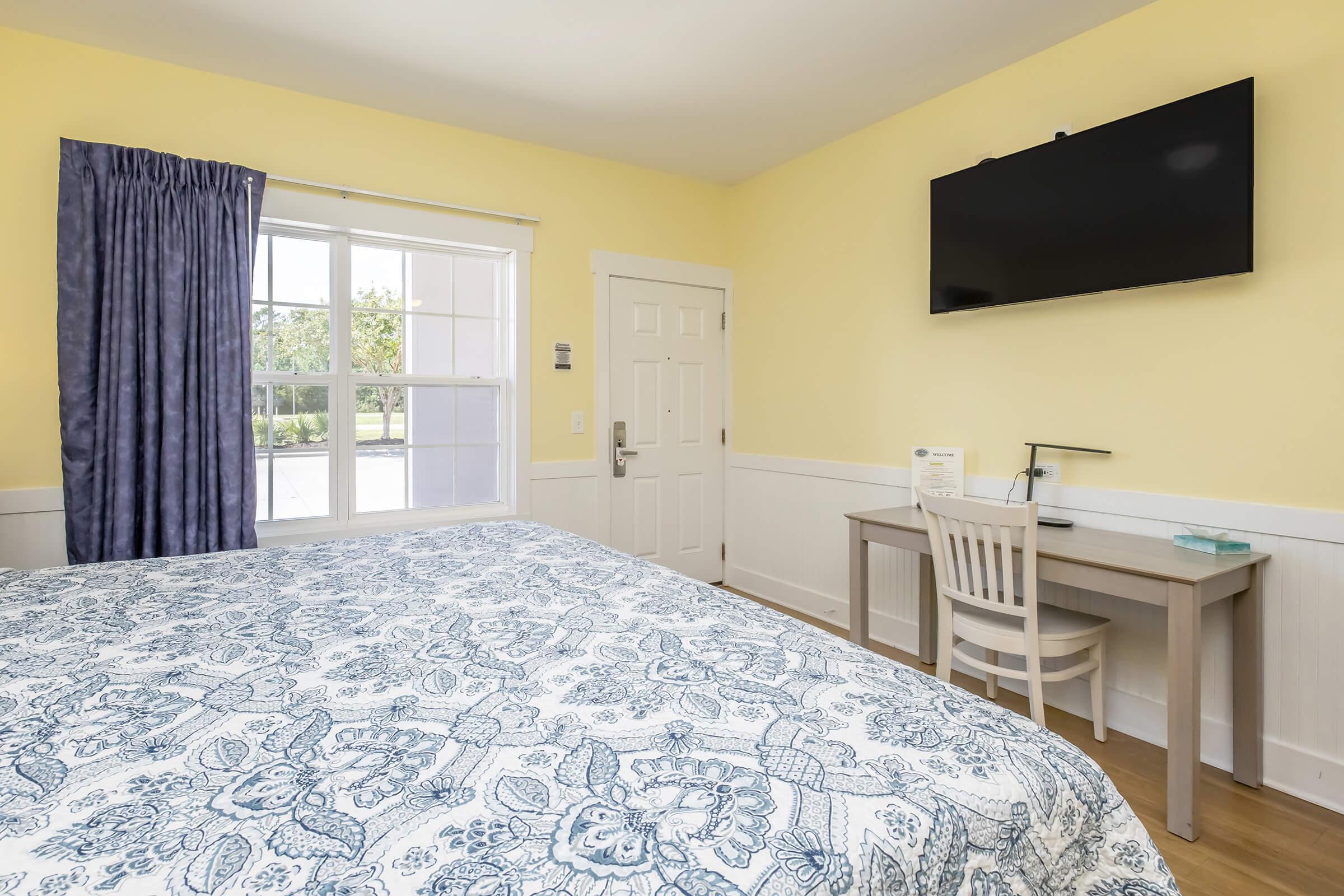 A brightly lit hotel room featuring a large window with sheer curtains, a bed with a patterned blue and white coverlet, a desk with a chair, and a mounted flat-screen TV on the yellow wall. The room has a cozy and inviting ambiance, showcasing a clean and organized space.