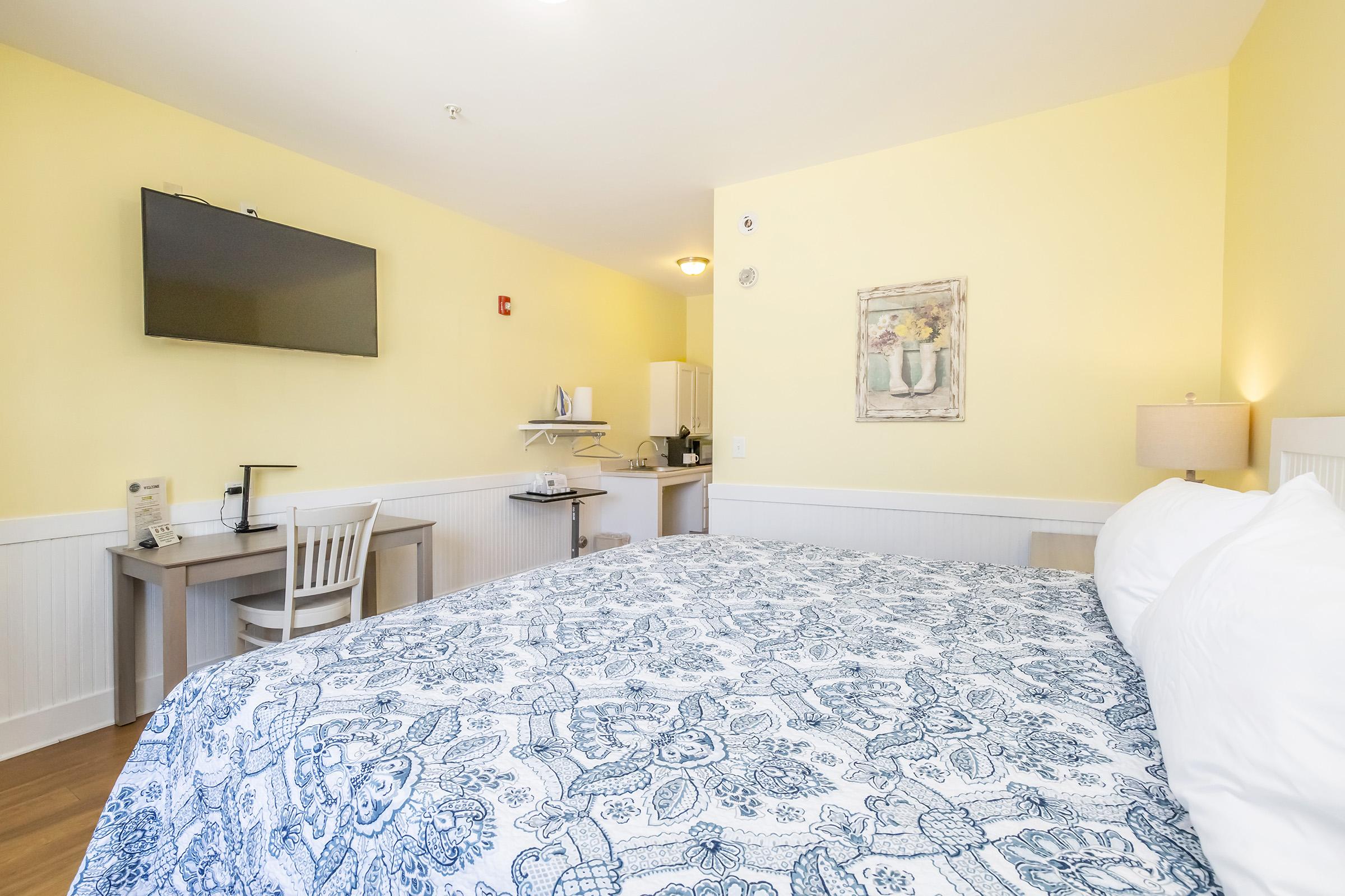 A cozy hotel room featuring a large bed with a patterned blue bedspread, a small wooden desk with a chair, and a flat-screen TV mounted on the wall. In the background, there is a kitchenette area with basic amenities, along with a decorative painting on the wall and a warm yellow color scheme.