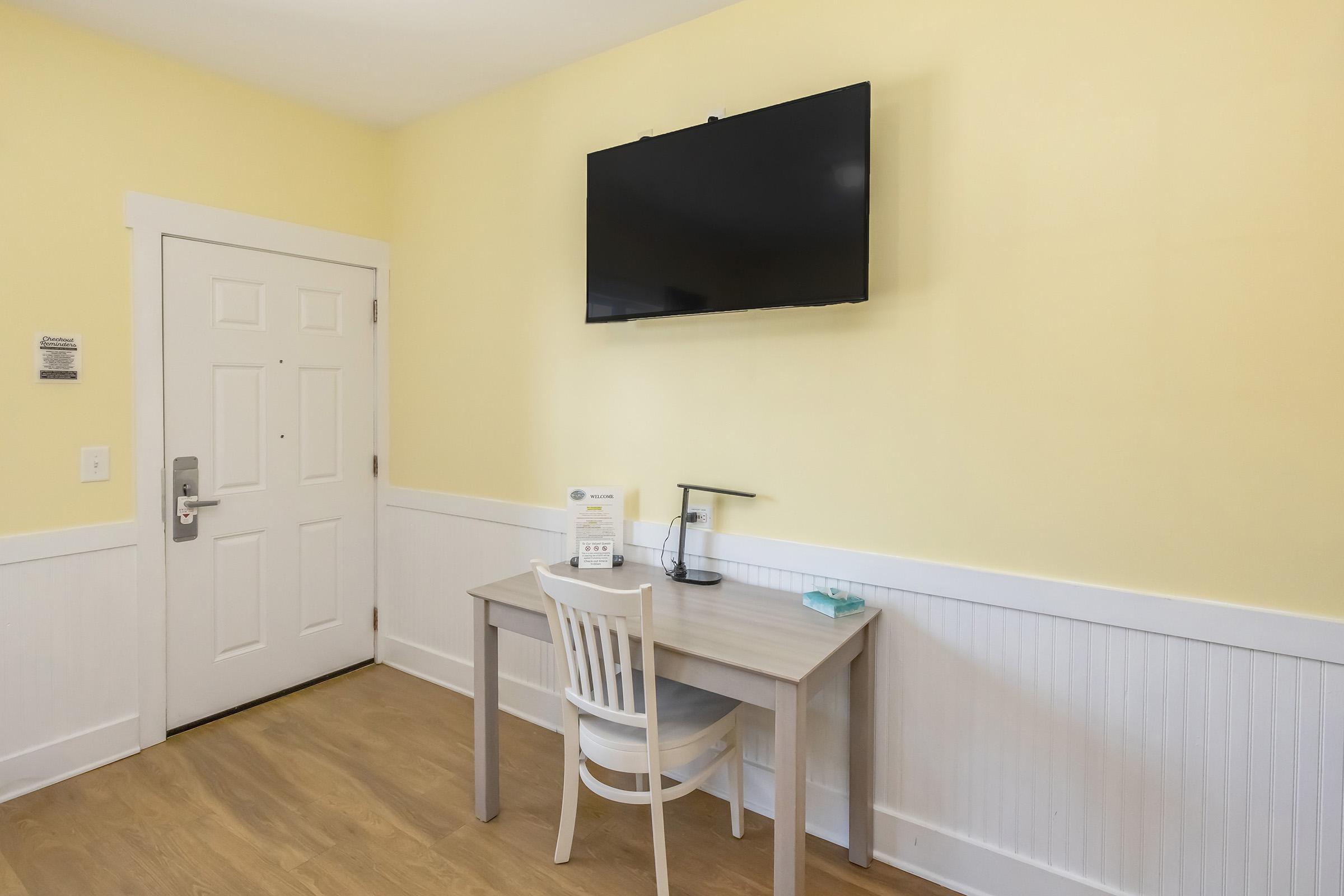 A simple room with yellow walls featuring a wooden desk and a white chair. A flat-screen TV is mounted on the wall, and there's a door visible on the left. The desk includes a lamp and a small blue item, adding to the minimalist decor.