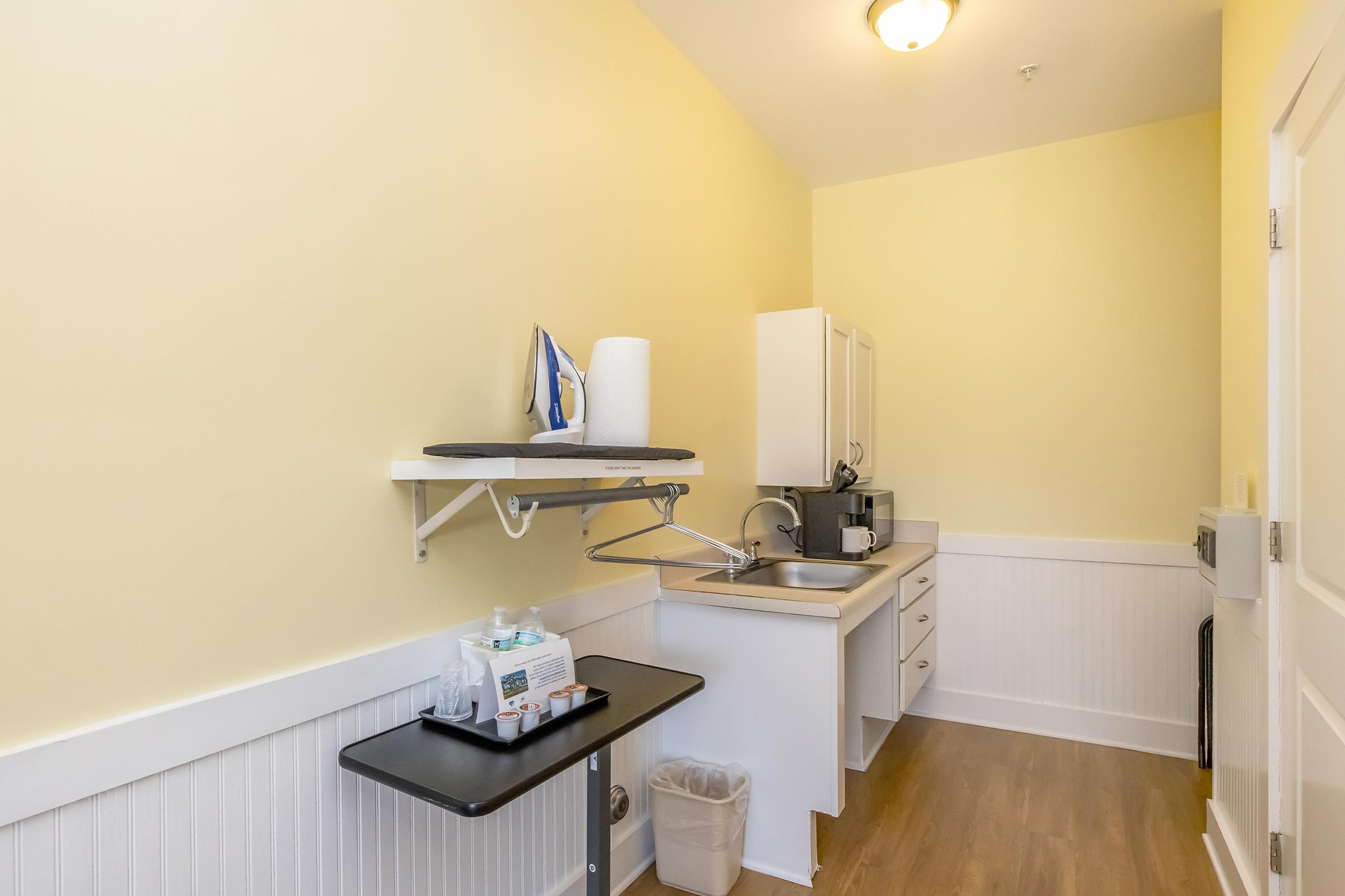 Small kitchenette area featuring a white sink, countertop, and microwave. There is a wall-mounted ironing board and an iron. A tabletop with supplies is on the left, next to a trash bin. The walls are painted light yellow, and the flooring is hardwood.