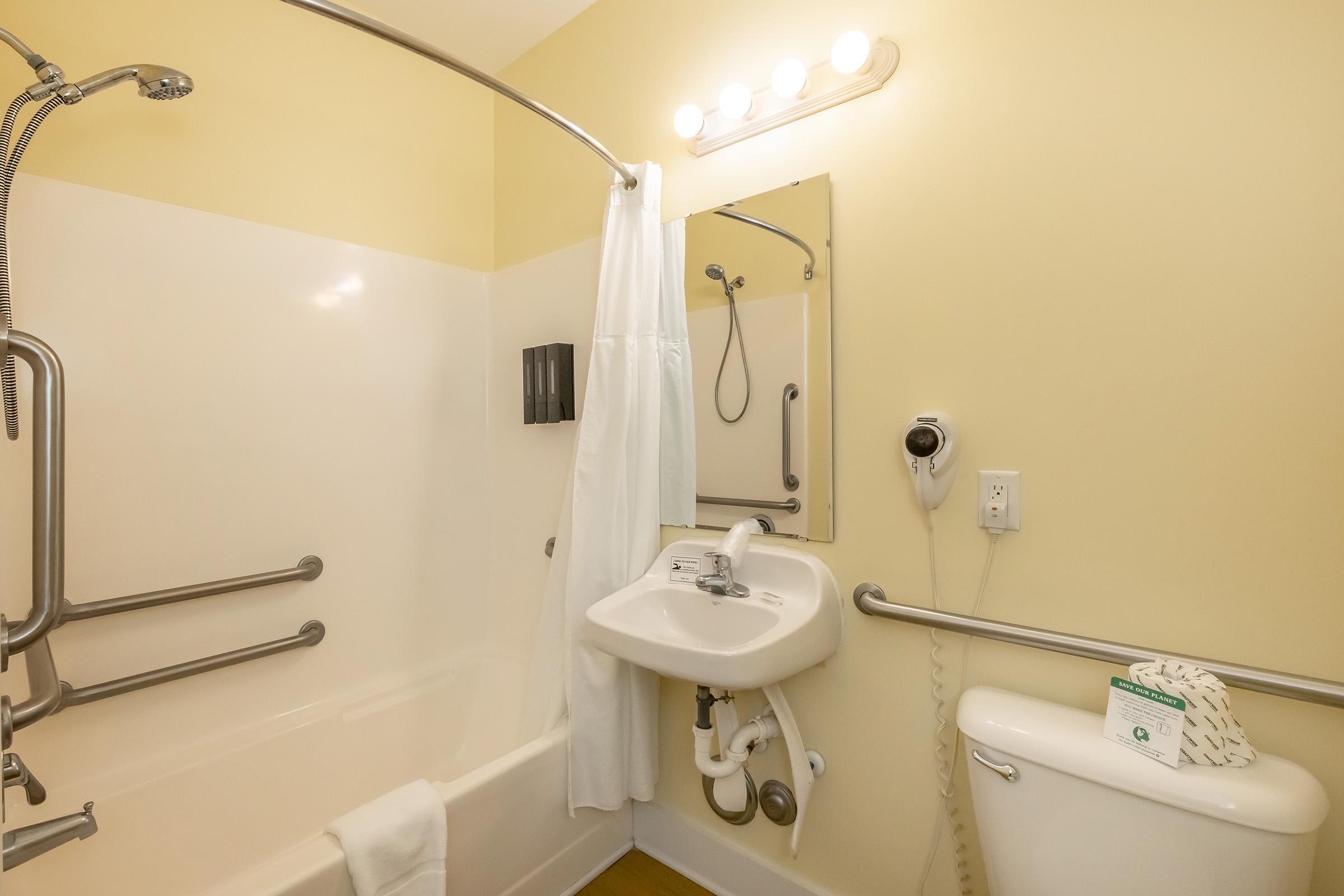 A clean, accessible bathroom featuring a bathtub with grab bars, a showerhead mounted on a wall, a mirror, a small sink, and a toilet. The walls are painted yellow, and there are safety features like handrails and a phone on the wall, enhancing usability for individuals with mobility challenges.