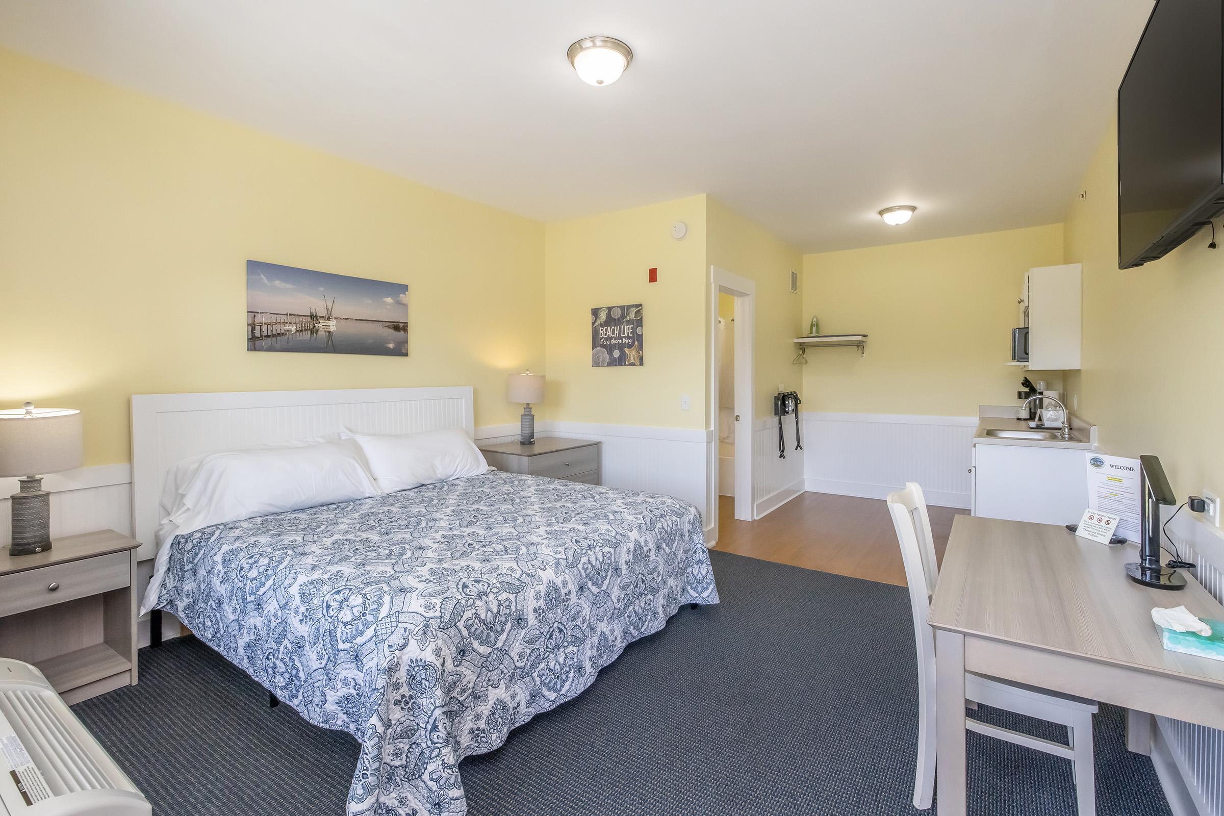 A cozy hotel room featuring a queen-sized bed with a blue and white patterned blanket, a bedside table with lamps, a work desk, and a small kitchenette. Light yellow walls create a warm atmosphere, and there is artwork on the wall depicting a pier scene. A flat-screen TV is mounted on the wall, and the flooring is carpeted.