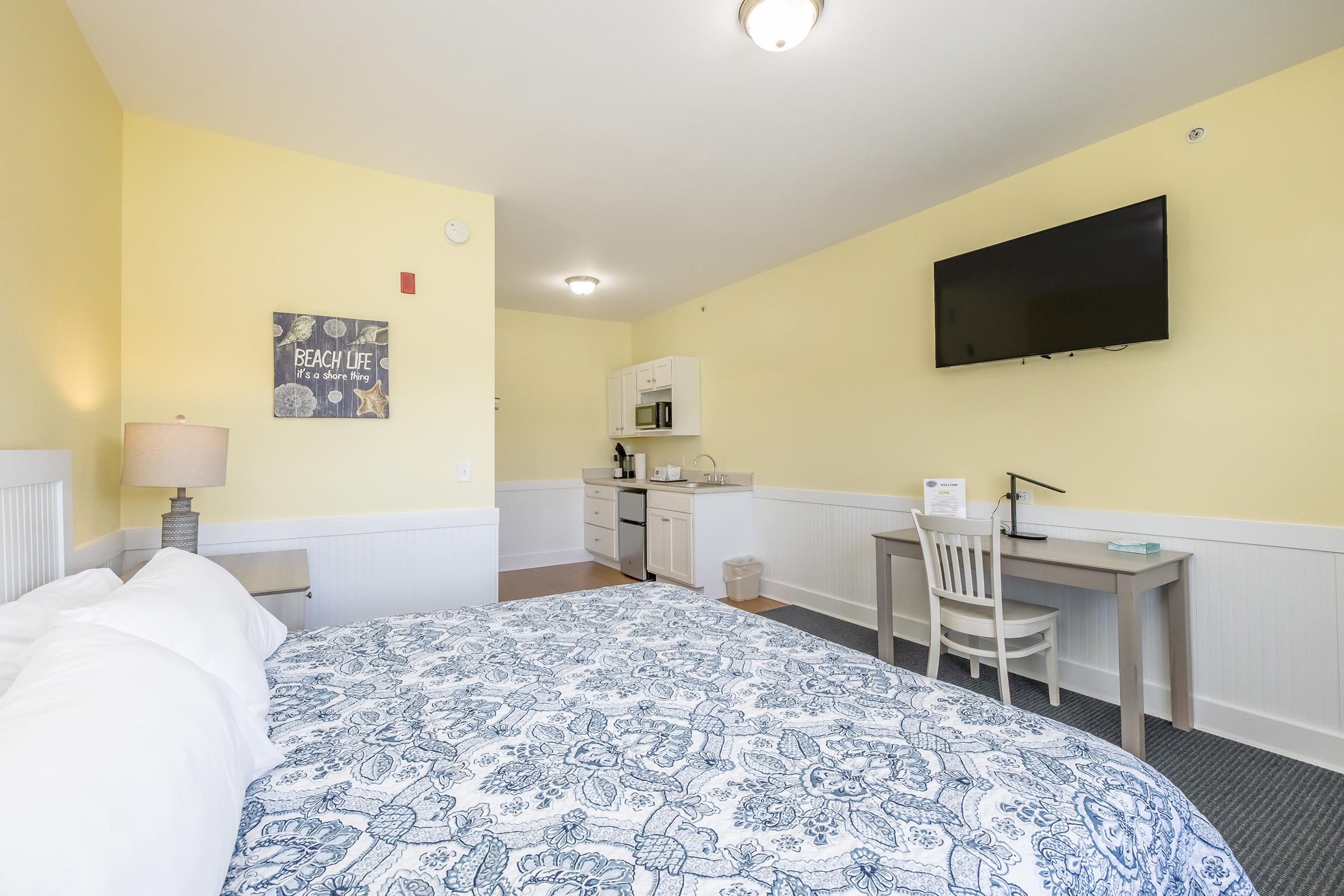 A bright, inviting hotel room features a large bed with a blue and white patterned quilt. There's a small kitchenette area with a microwave and refrigerator. A desk with a chair is positioned near a wall-mounted TV. The walls are painted yellow, and a decorative sign reading "Beach Life" is displayed.