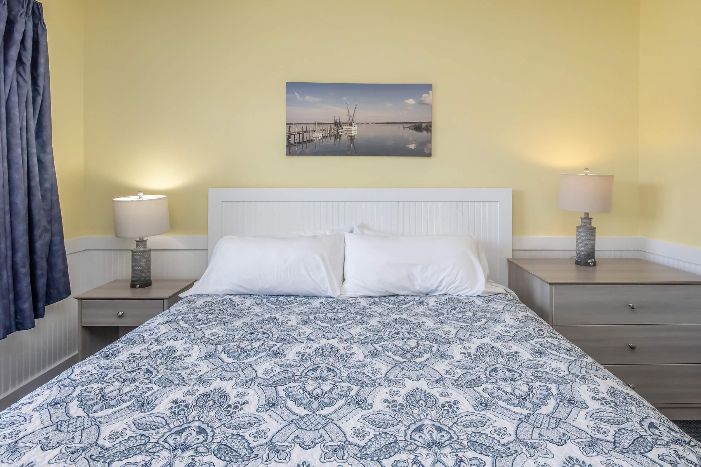 A cozy bedroom featuring a neatly made bed with white pillows and a blue patterned blanket. There are light wooden nightstands on either side with lamps, and a scenic photograph of a pier and water hangs on the yellow wall. Natural light brightens the space through a curtained window.