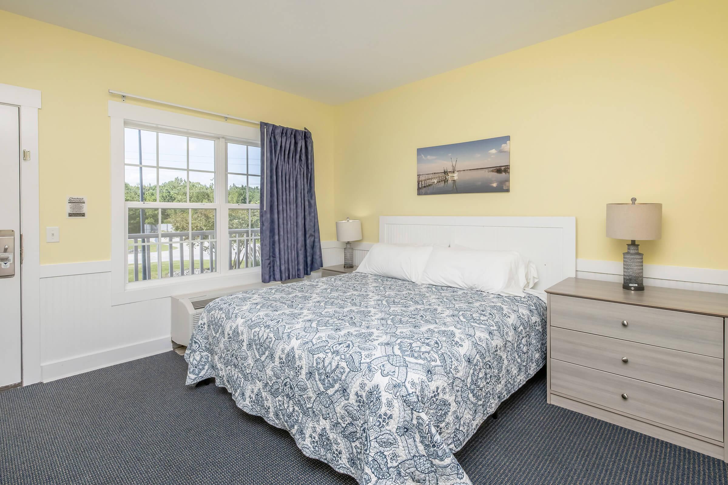 A cozy hotel room featuring a queen-sized bed with a blue and white patterned blanket. There's a bedside table with a lamp beside the bed, a large window dressed with blue curtains, and a small artwork on the wall. The room has light yellow walls and a carpeted floor. A dresser is also visible in the room.