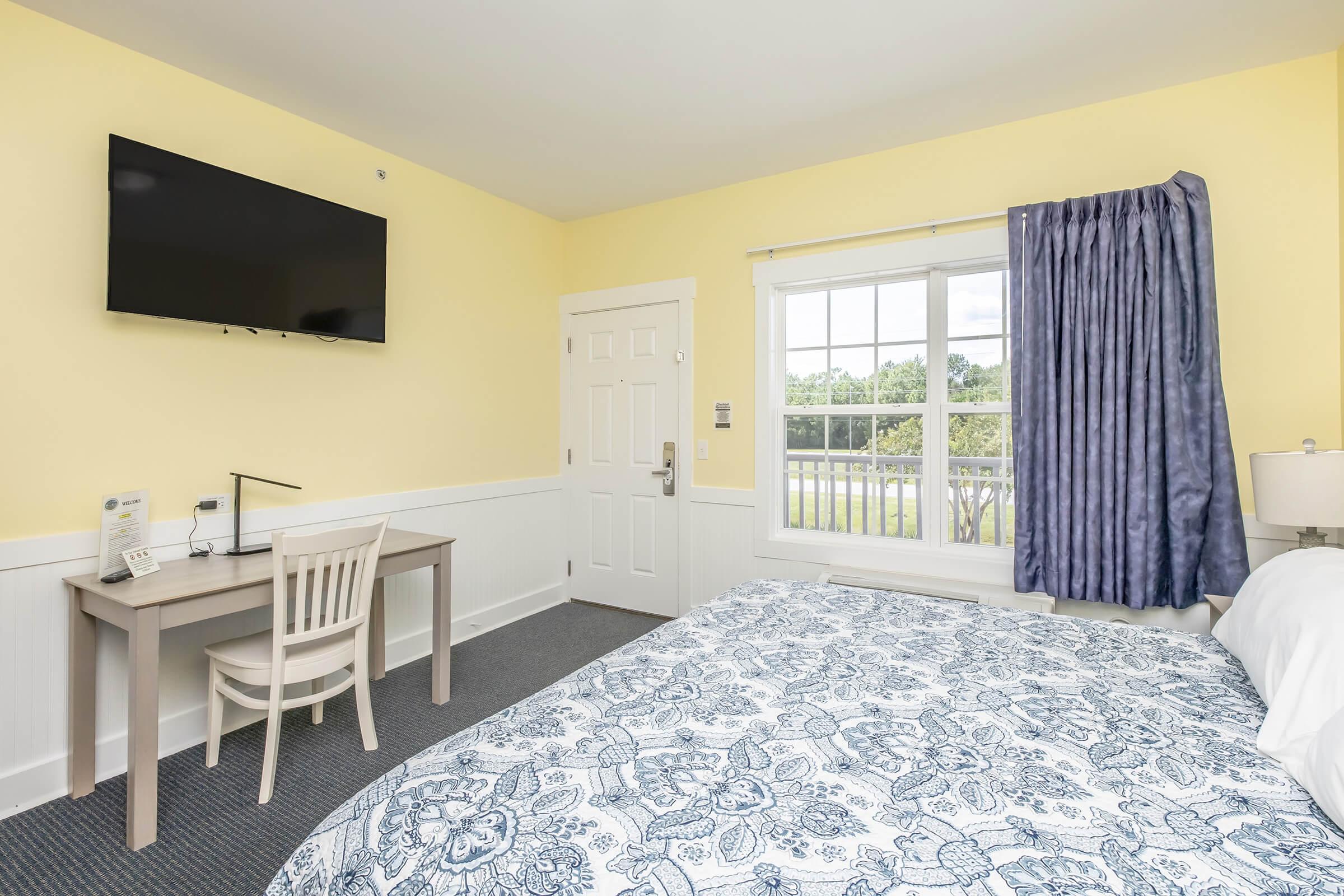 A bright and cozy hotel room featuring a bed with a blue patterned comforter, a desk with a chair, and a flat-screen TV mounted on the wall. The room has large windows with purple curtains, letting in natural light, and is decorated with soft yellow walls.