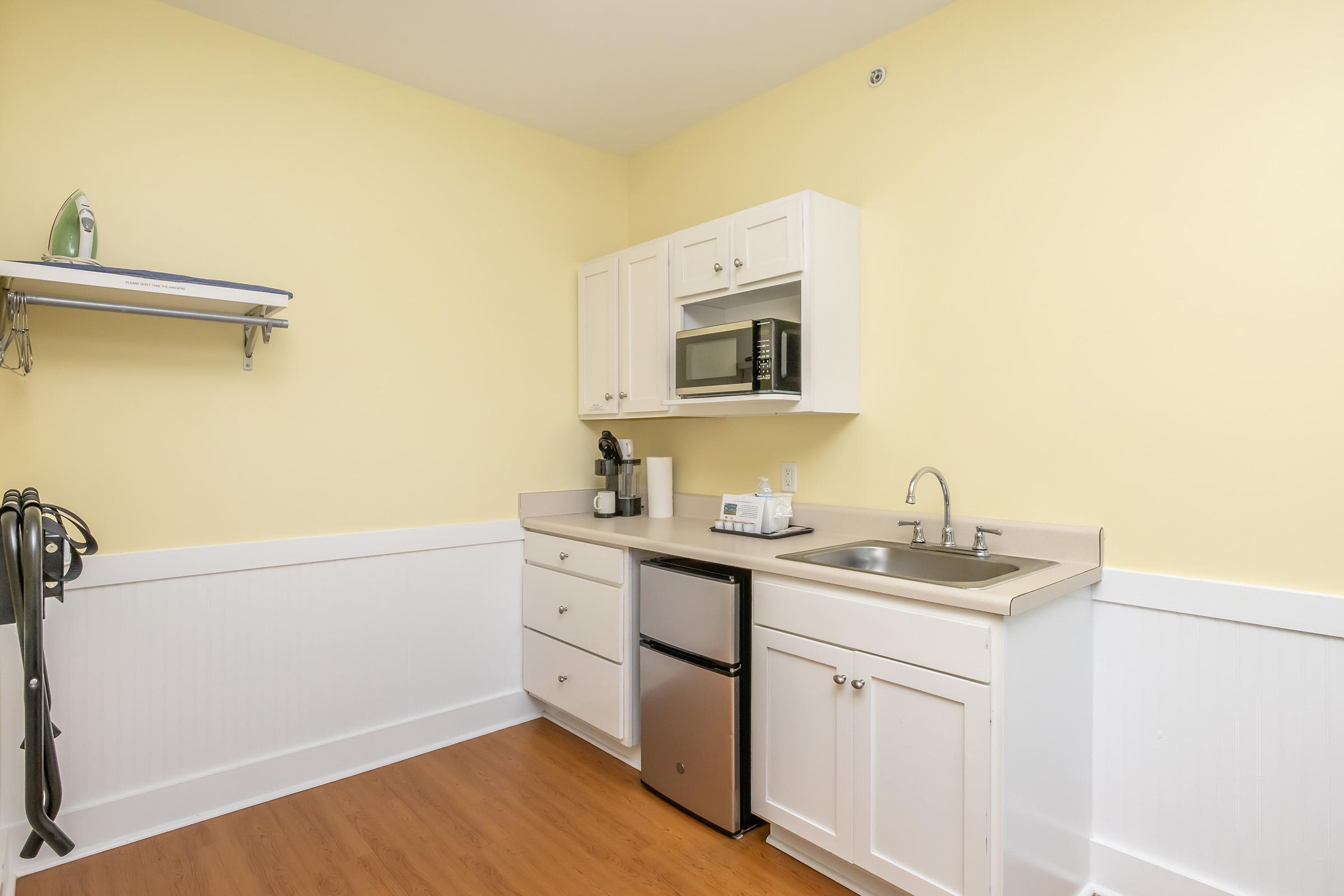 A small kitchen area with yellow walls, featuring white cabinets, a stainless steel sink, a microwave, a coffee maker, and a compact refrigerator. There is a shelf above the countertop and a light wooden floor.