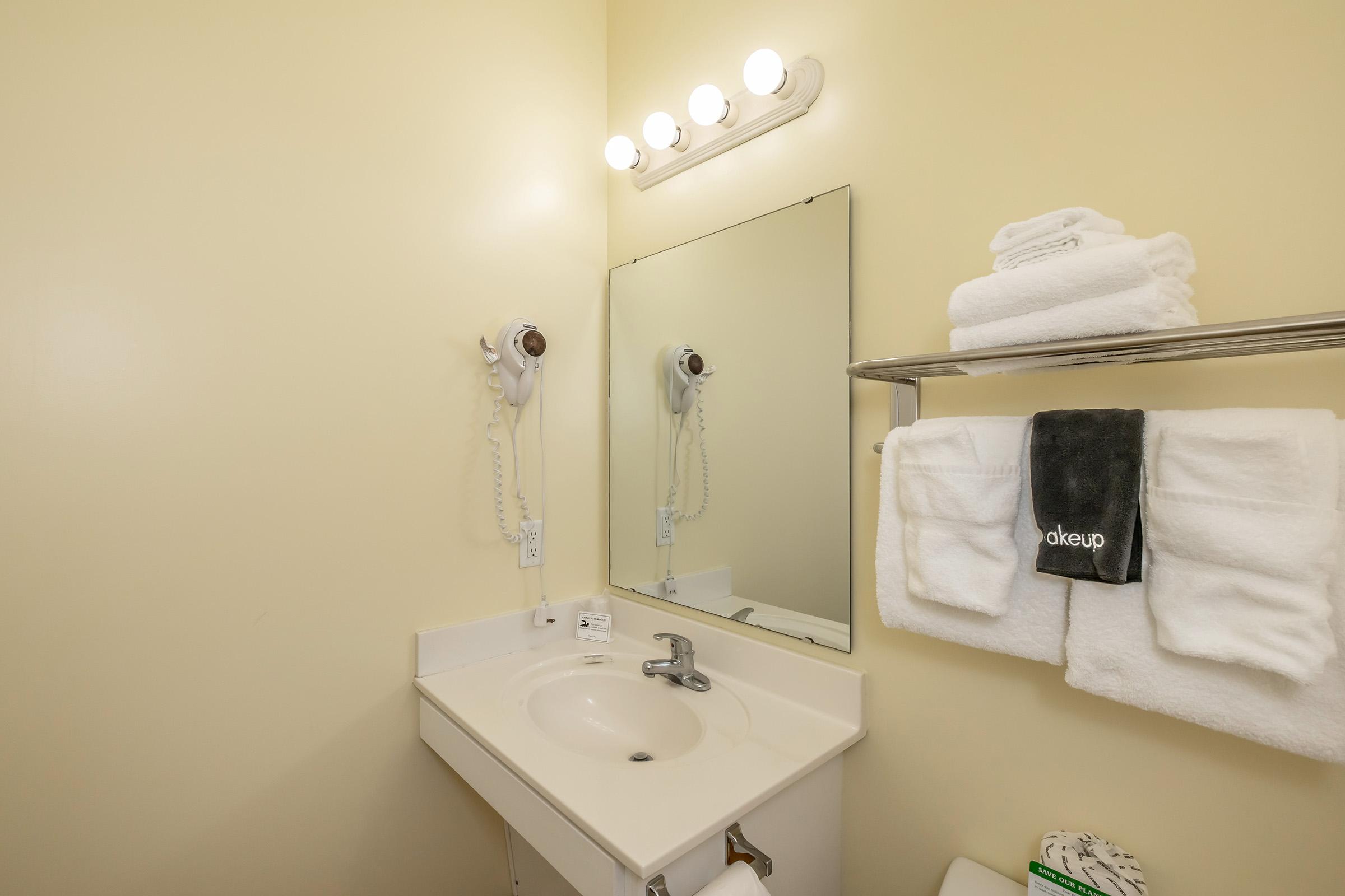 A small bathroom featuring a white sink, a mirror above it, and a wall-mounted hairdryer. Soft, stacked white towels are neatly arranged on a rack next to the sink. There are light fixtures above the mirror, and the walls are painted in a light yellow color.