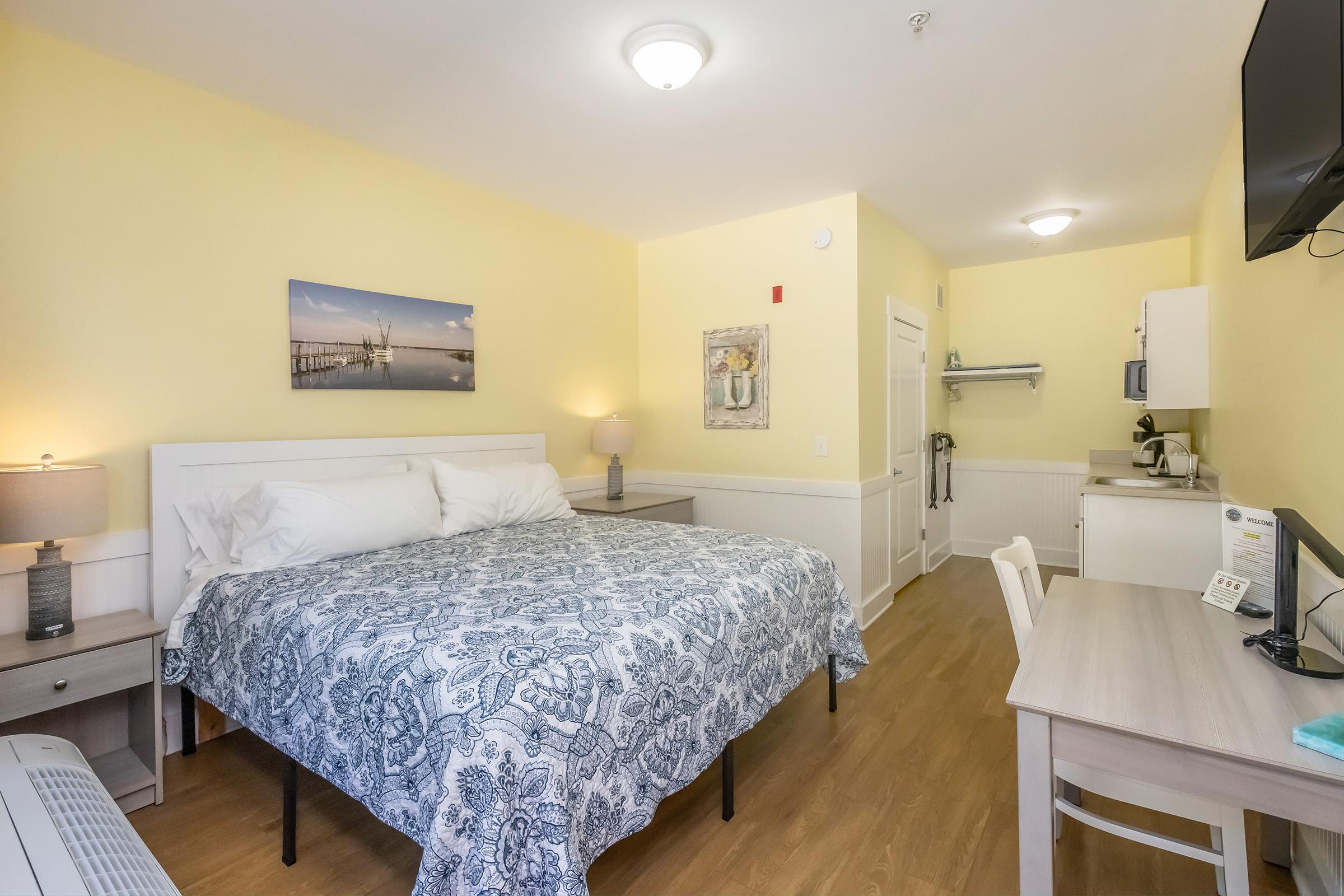 A cozy hotel room featuring a queen-sized bed with a patterned blue and white bedspread. The room is painted in soft yellow, with a desk, chair, and flat-screen TV. There are lamps on side tables and a kitchenette area visible in the background, enhancing the inviting and comfortable atmosphere.