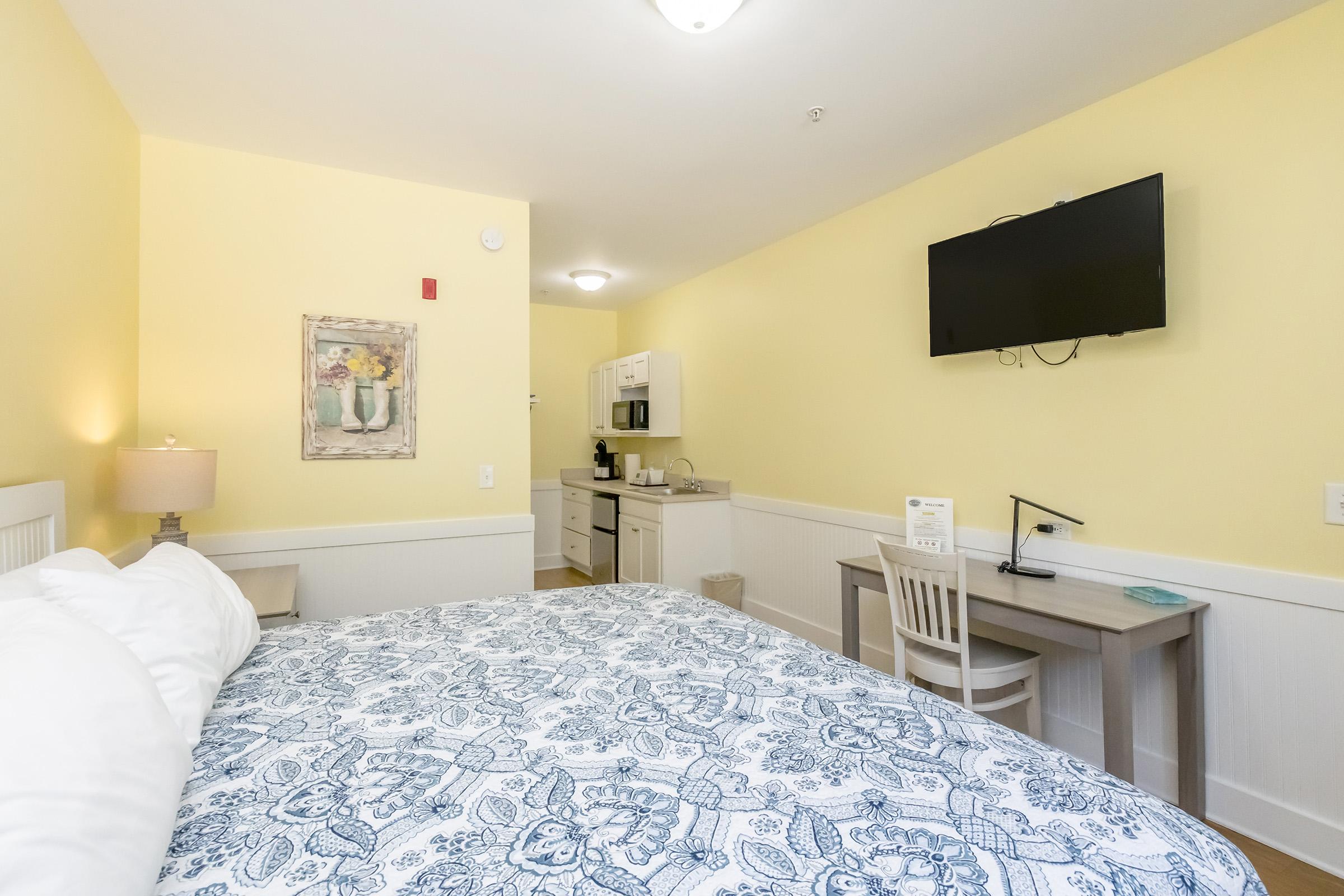 A cozy hotel room featuring a large bed with a patterned blue and white duvet, a desk with a chair, and a flat-screen TV mounted on the wall. The room has yellow walls, a kitchenette with a microwave, and a small table. A piece of artwork hangs above the bed, adding a decorative touch.