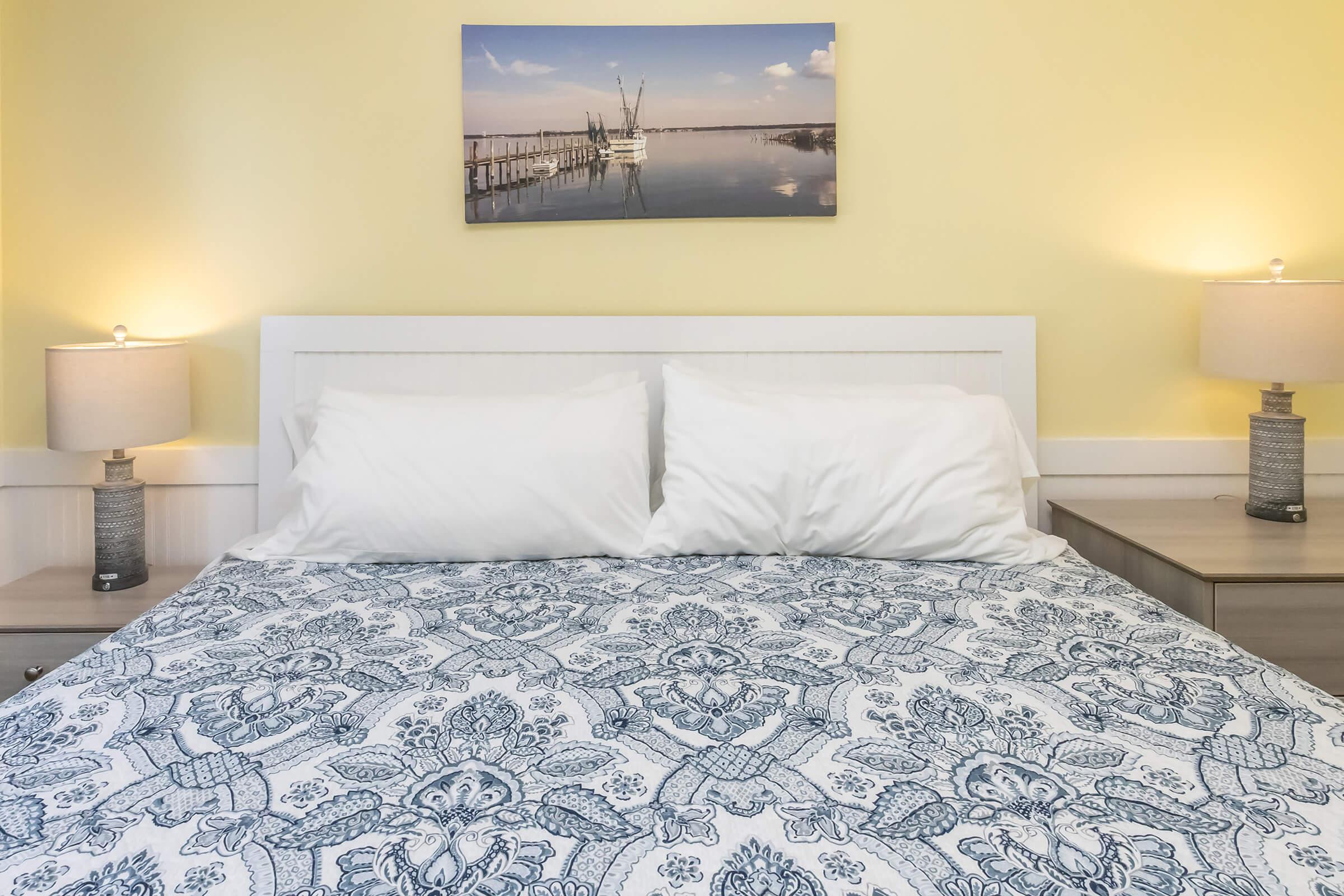 A cozy bedroom featuring a white bed frame with two fluffy pillows and a decorative blue patterned bedspread. On either side of the bed are small nightstands with stylish lamps. Above the bed hangs a picture of a serene waterfront scene, enhancing the calming atmosphere of the room.