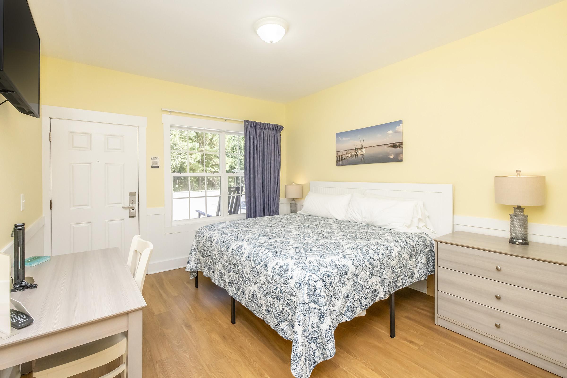 A bright and inviting hotel room featuring a queen-sized bed with a blue and white patterned bedspread, a wooden nightstand with a lamp, a dresser, and a work desk. A window allows natural light, and a flat-screen TV is mounted on the wall. The walls are painted a cheerful yellow, creating a warm atmosphere.