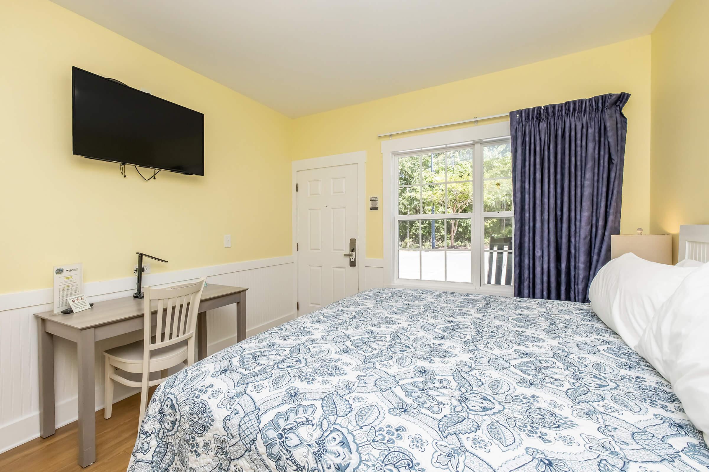 A cozy room with a queen-sized bed featuring a blue and white patterned duvet, a small desk and chair, and a wall-mounted TV. Natural light comes through large windows covered with dark curtains, enhancing the warm yellow walls and wooden flooring. A door leads outside, adding to the inviting atmosphere.