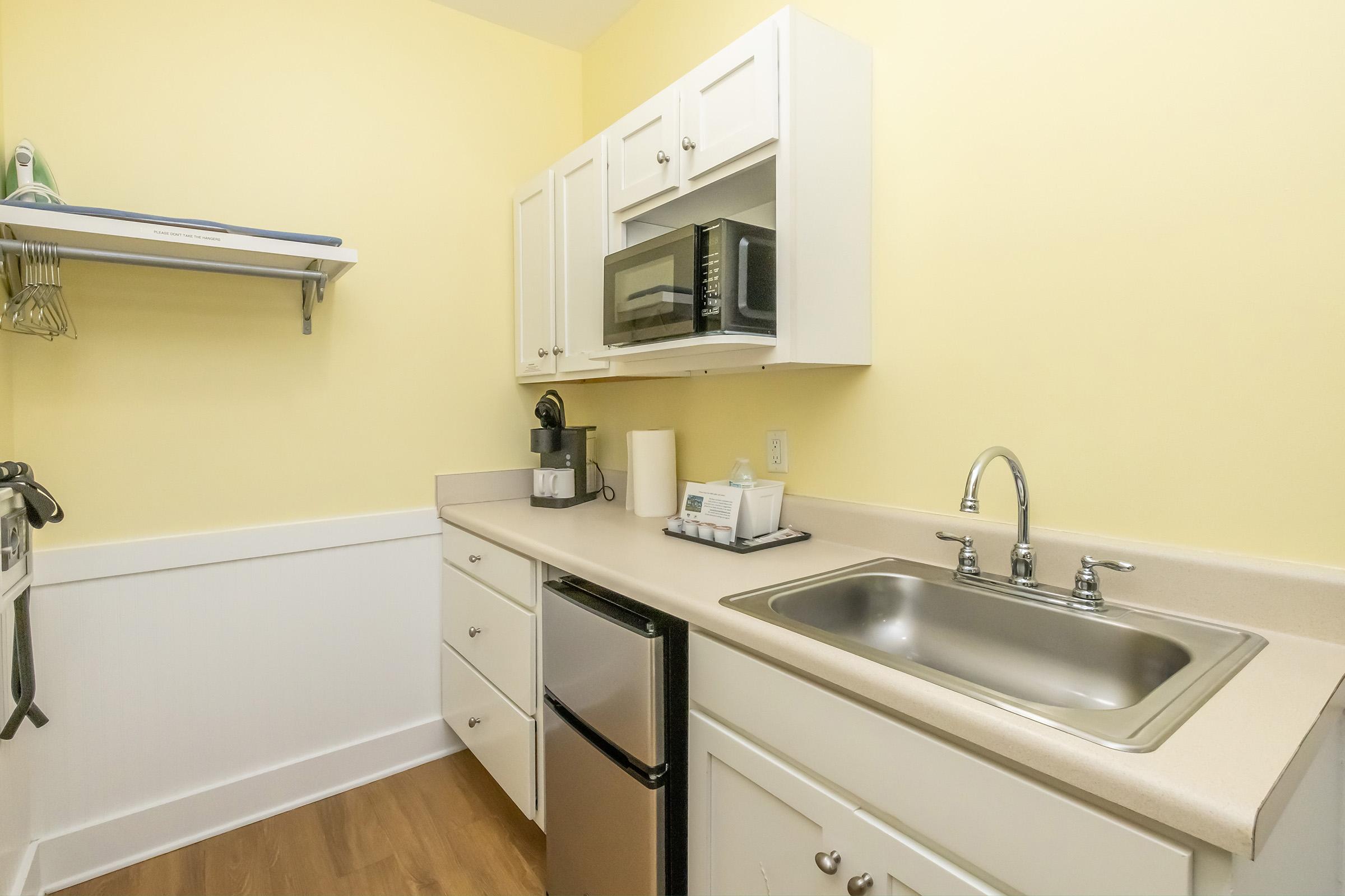 A small kitchen with yellow walls. It includes white cabinets, a stainless steel sink, a microwave, a coffee maker, and a compact refrigerator. There's a countertop for food preparation, and an iron and ironing board are mounted on the wall, providing a tidy and functional space.