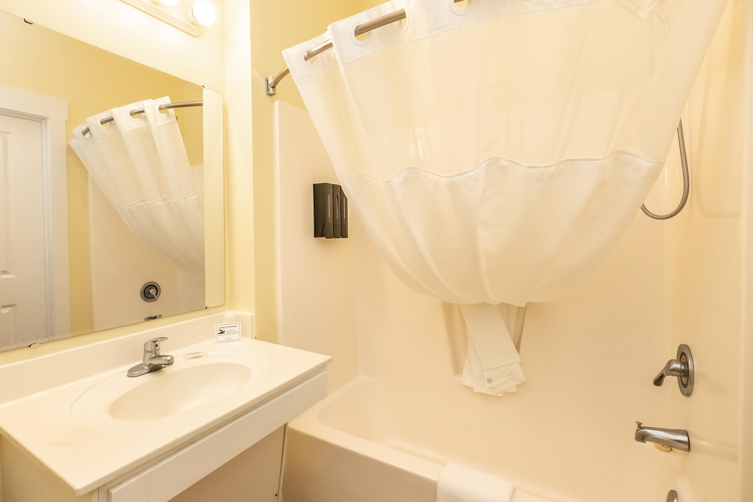 A small bathroom featuring a beige tub and shower, with a white shower curtain hanging. There is a mirror above a white sink with a faucet, and a towel hanging nearby. The walls are painted a light yellow, creating a bright and clean atmosphere.