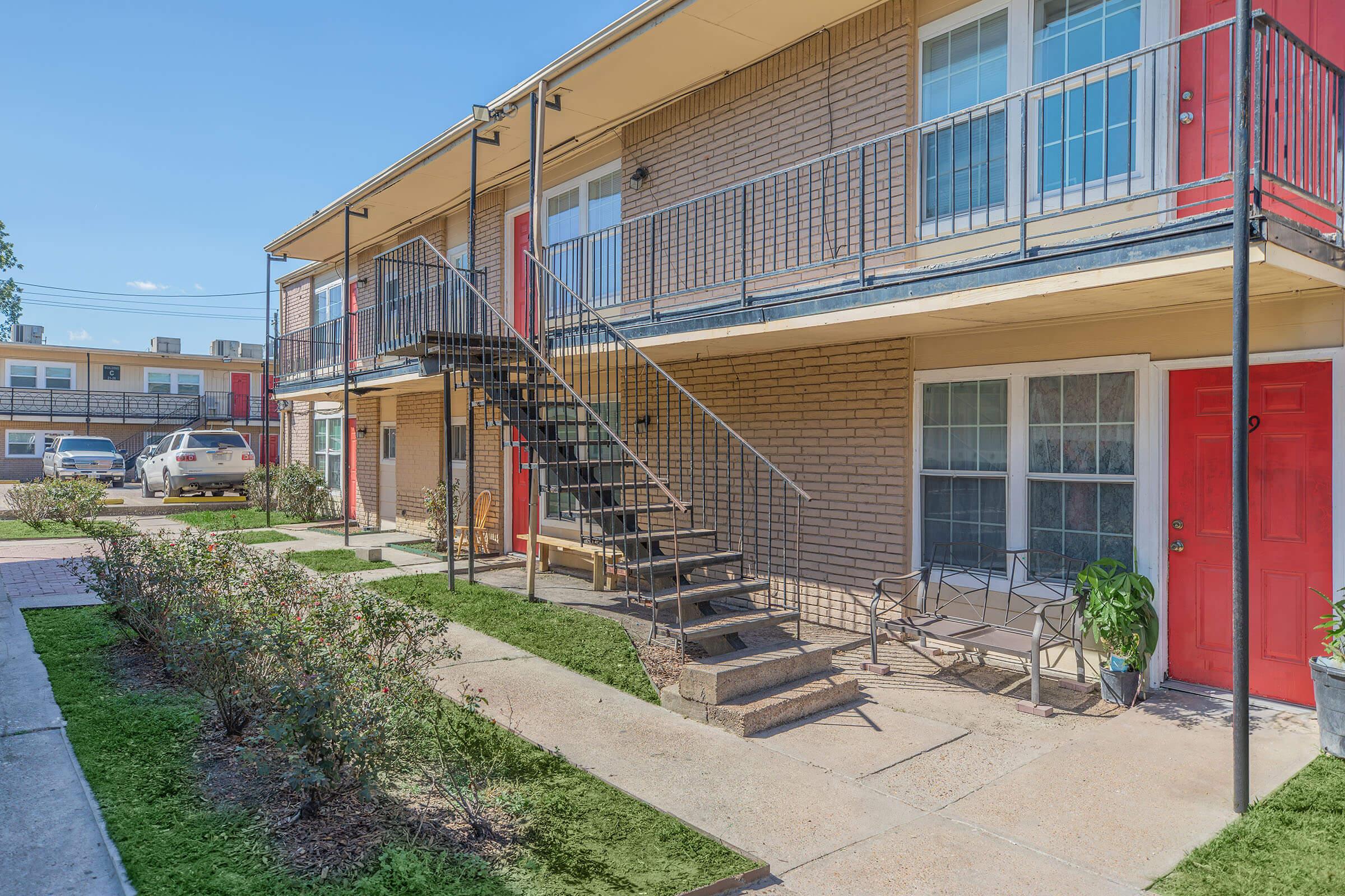 A view of a two-story apartment building featuring metal staircases, red doors, and a small landscaped courtyard. Pathways lead to individual entrances, and a parked car is visible in the background. The setting appears well-maintained with grass and plants along the walkway.