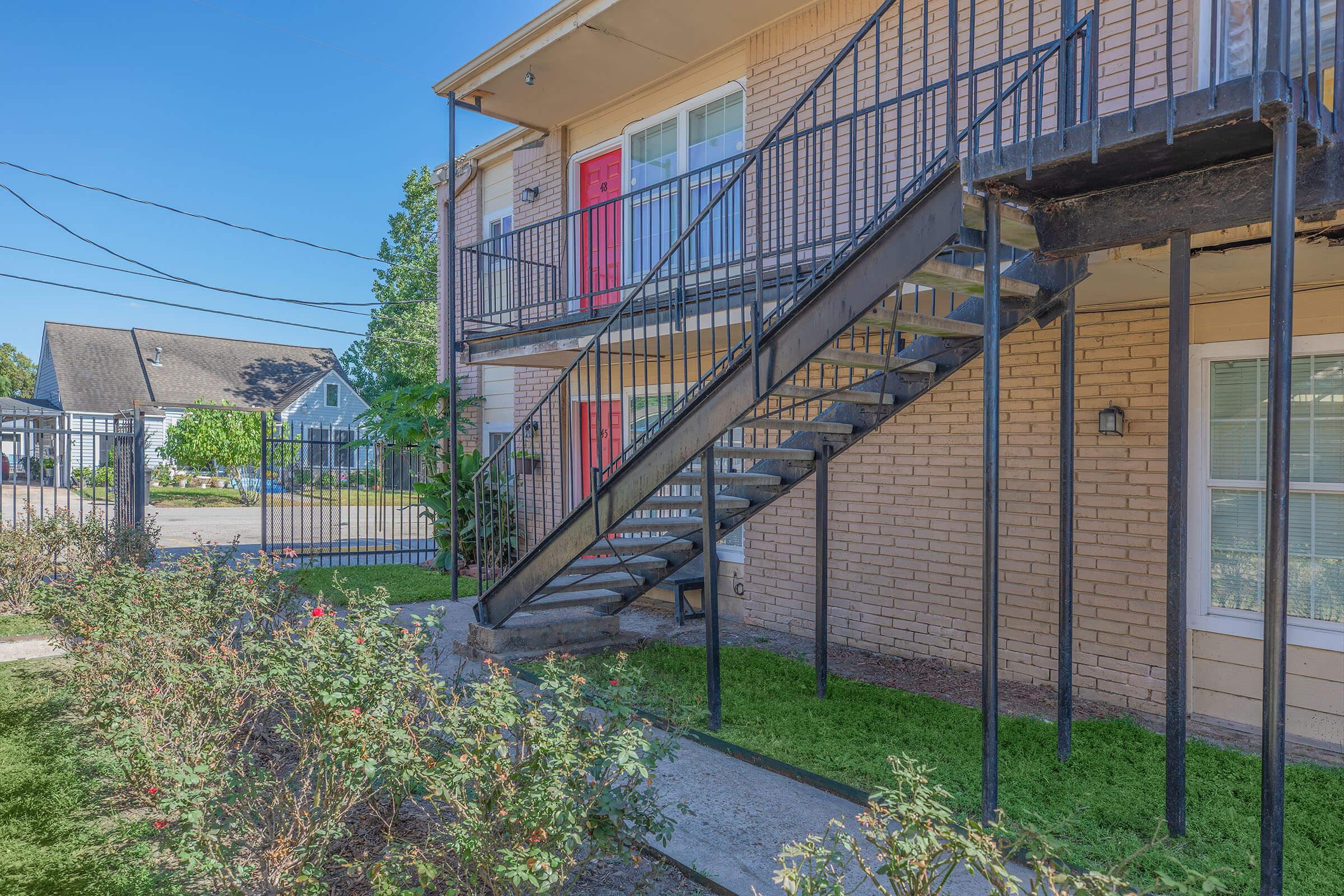 A view of a two-story apartment building featuring a red front door, outdoor staircase, and a small garden area with blooming flowers. The building is surrounded by a fence, and utility wires are visible in the background under a clear blue sky.