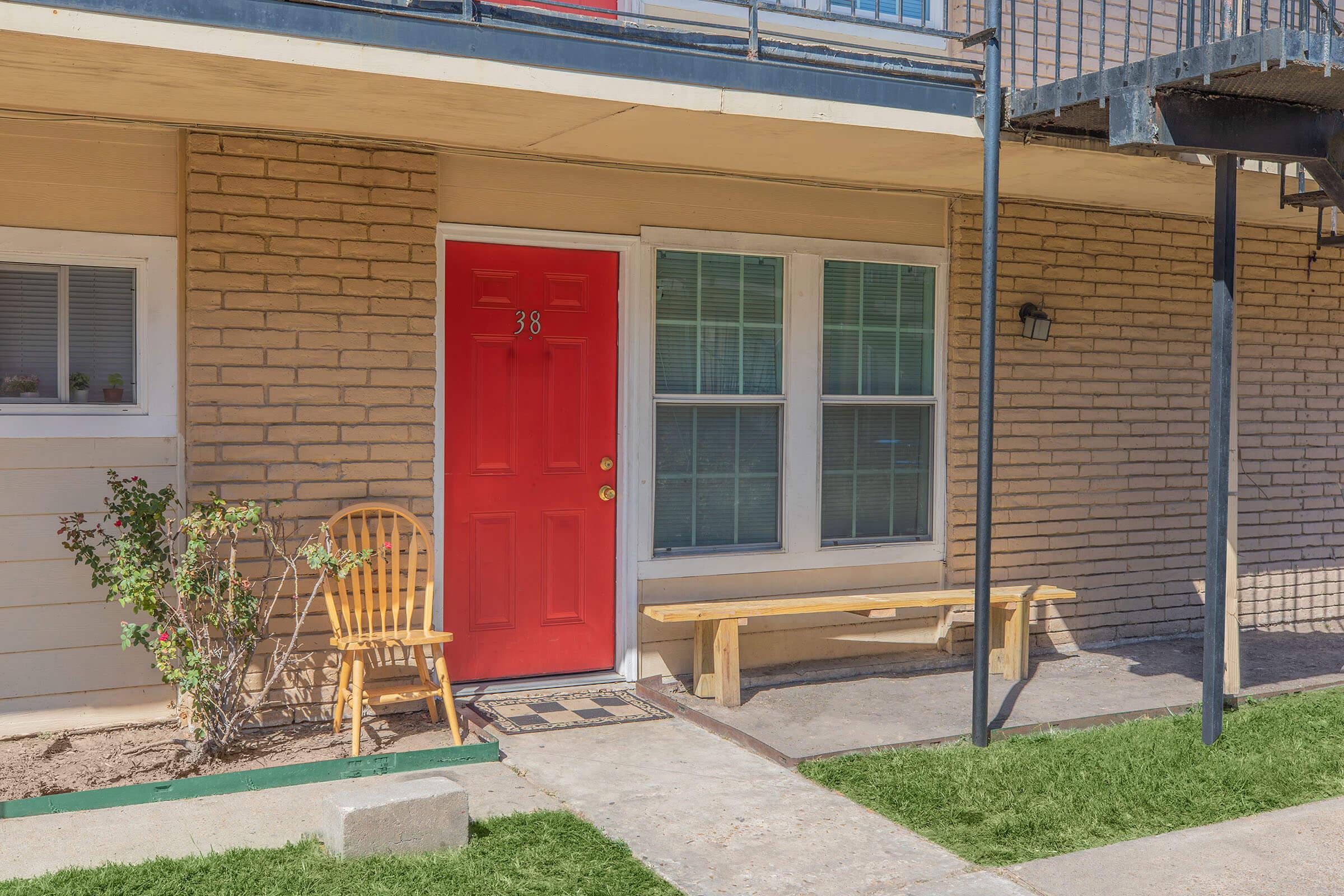 A ground-level doorway with a bright red door marked "38." To the left, a wooden chair and a bench are placed on a concrete pathway. The exterior features beige brick walls and a small window with greenery visible. A metal staircase leads up to the next level.