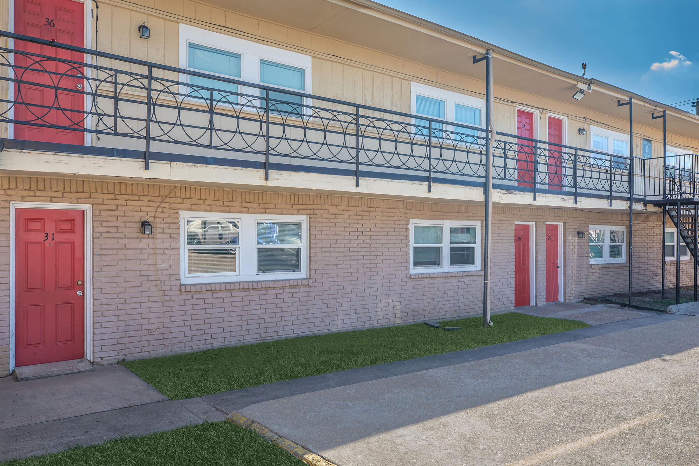 Two-story motel with several rooms; the upper floor features rooms with red doors and a black railing, while the lower level has similar rooms with an adjoining pathway. The ground is covered with green grass, and there is a visible concrete pathway leading to the entrance. Bright blue sky above.
