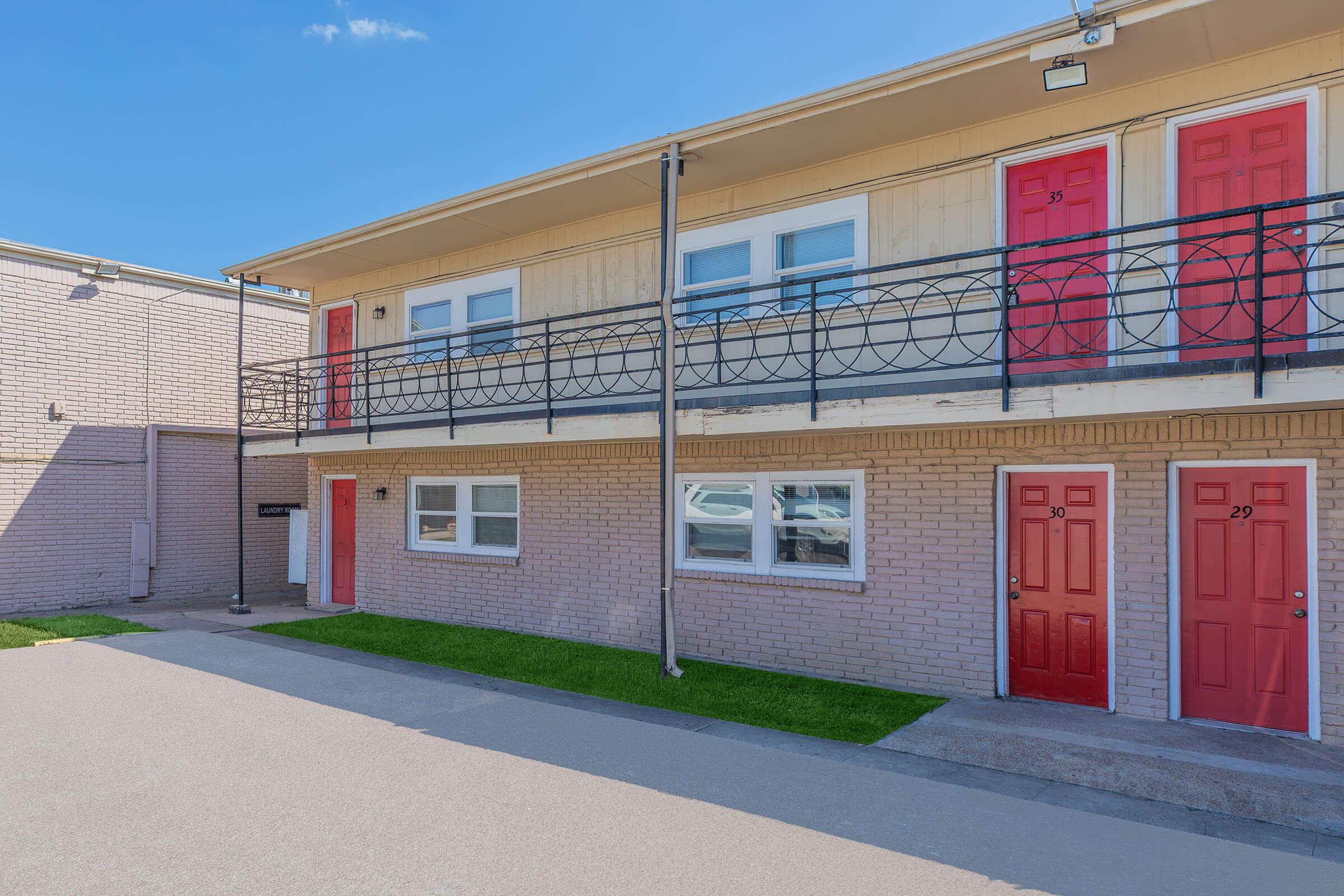 A view of a two-story motel building featuring red doors and a balcony. The ground level has multiple doors labeled with numbers 29 and 30. The exterior is a light-colored brick with a gray pathway leading up to the entrance. A clear blue sky is visible above, and the surroundings are well-maintained grass.