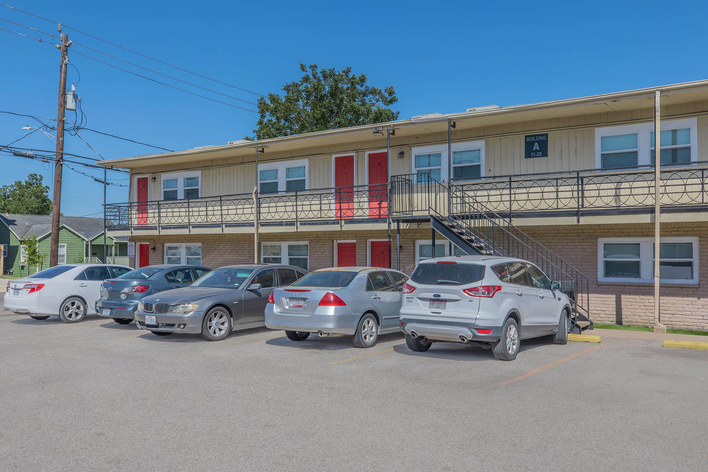 Two-story motel-style building with a beige exterior and red doors. A set of stairs leads to the second floor. Several parked cars are lined up in front, with clear blue skies above. The scene conveys a tidy, inviting atmosphere typical of a budget accommodation.