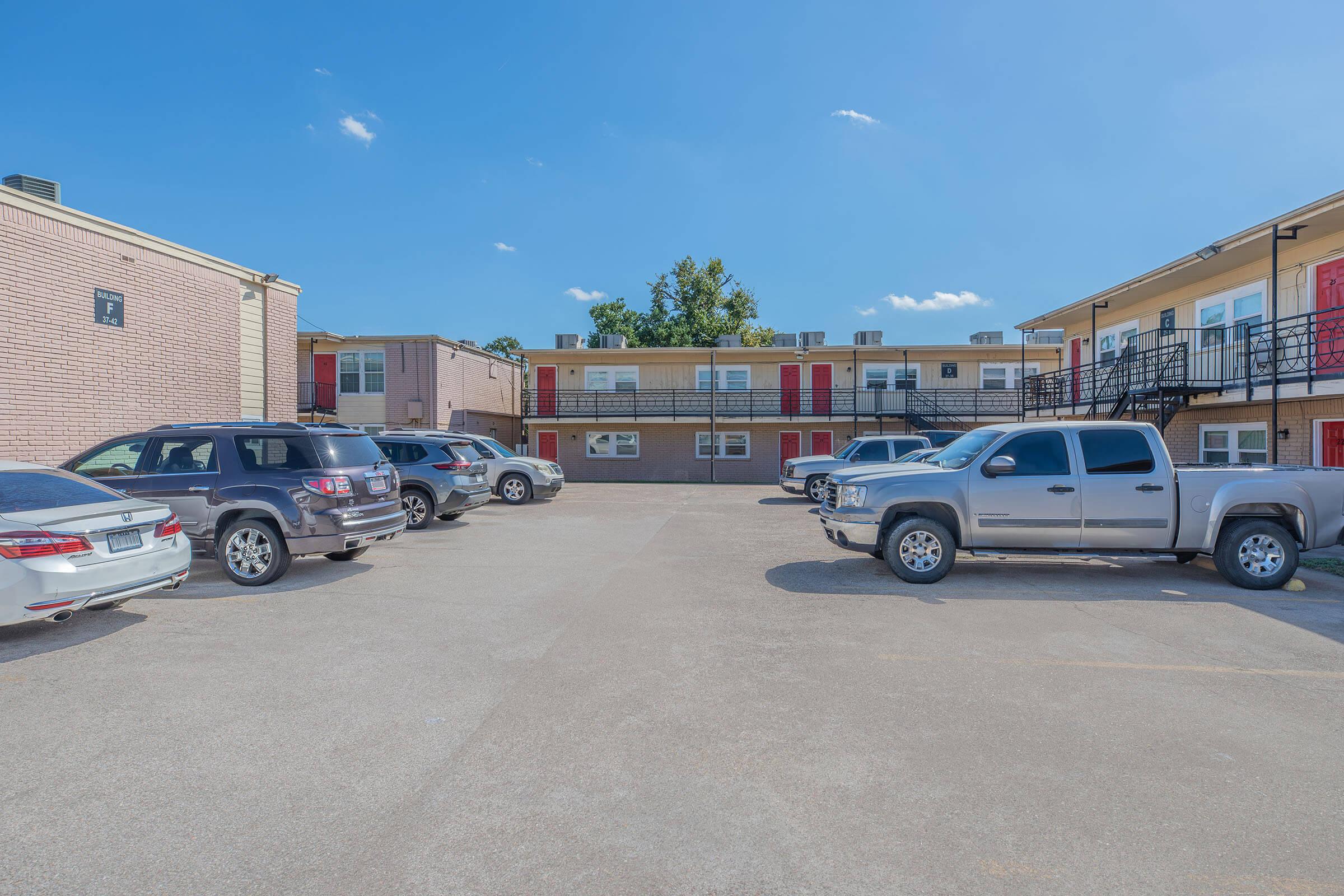 A parking lot with several cars parked in front of a two-story apartment building. The building features red doors and windows, surrounded by trees and a clear blue sky. The scene captures a sunny day in a residential area.