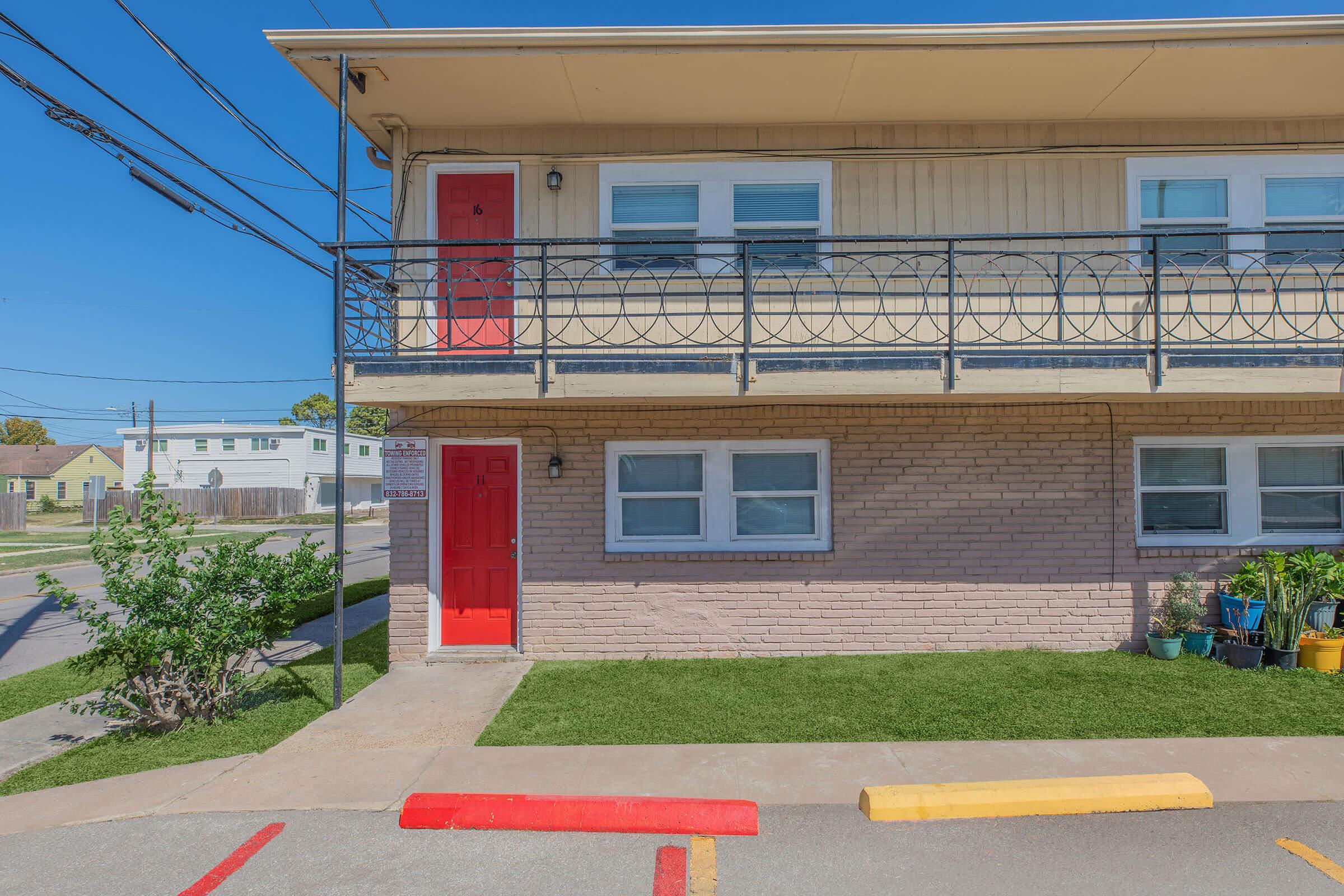 A two-story residential building with a light brown exterior and red doors. The balcony features a decorative railing, and there is a small green lawn in front. The driveway includes yellow and red painted sections. Power lines are visible in the blue sky.