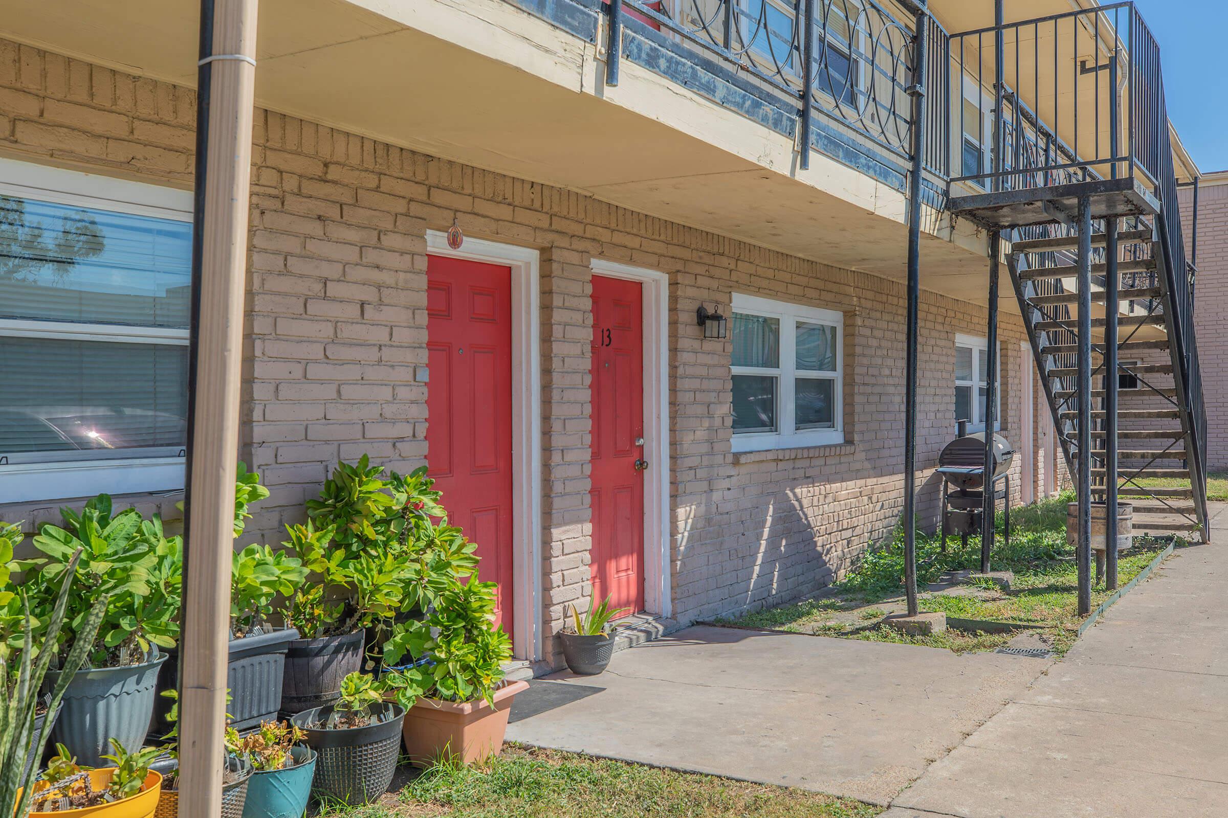 Two bright red doors on a light brick apartment building, with a balcony above and a metal staircase on the right. Potted plants are arranged along the walkway. The scene is sunny and well-maintained, showcasing a tidy outdoor space.
