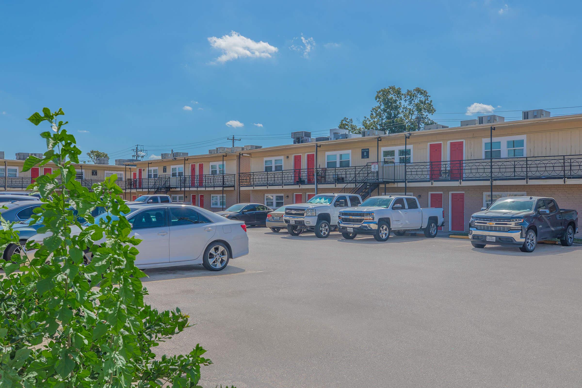 A parking lot in front of a two-story motel with red doors. Several cars are parked, and the building features a light color with black railings. The sky is clear with a few clouds. Green foliage is visible in the foreground.