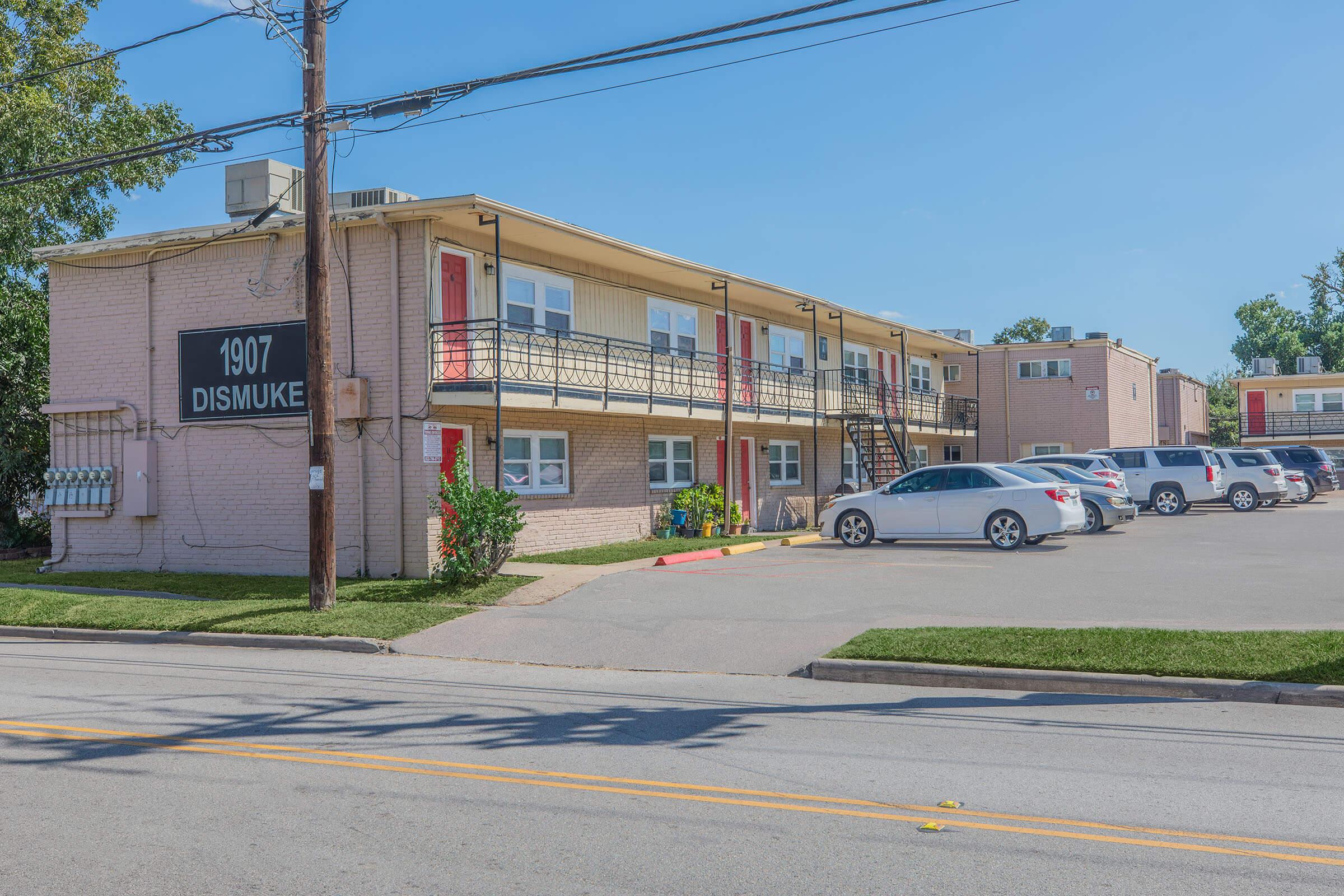 A view of a two-story apartment building with a light pink exterior, featuring multiple balconies and a black sign reading "1907 DISMUKE." There are several cars parked in front and a clear blue sky overhead. The setting is an urban area with trees in the background.