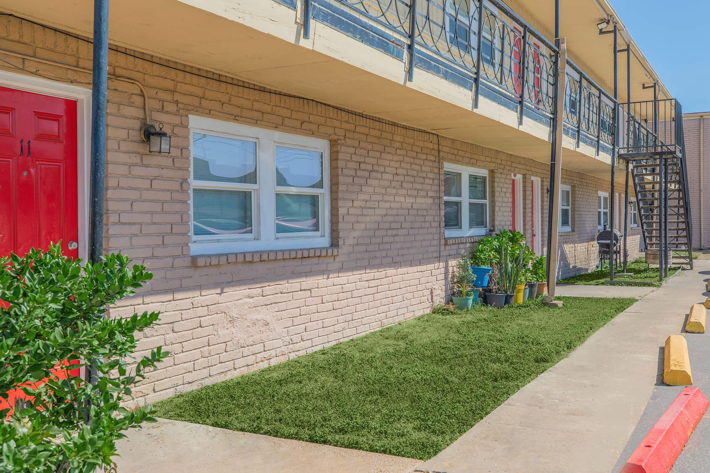 Exterior view of a one-story motel with a red door. The building features beige brick walls, white windows, and a balcony above with metal railings. There is a small patch of artificial grass and potted plants in front. A stairway leads to the upper level on the right side, with a parking area visible in the foreground.
